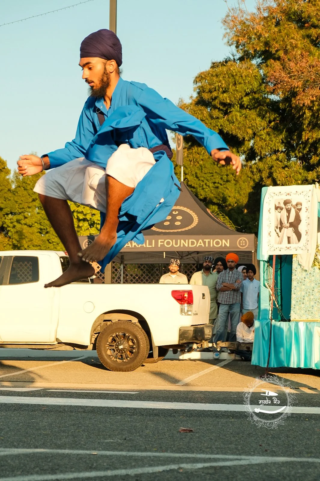 A man dressed in traditional Punjabi attire, including a blue kurta, white pajama, and turban, performing a jump in front of a small float during a parade, with a group of onlookers and a tent in the background.