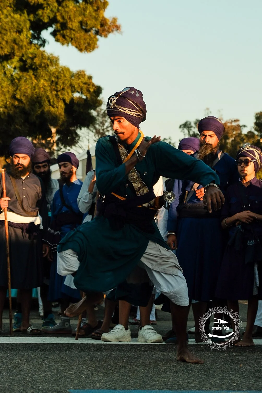 A man dressed in traditional Indian attire performing a street dance in front of a crowd of onlookers, all wearing turban-style head coverings, outdoors during sunset.