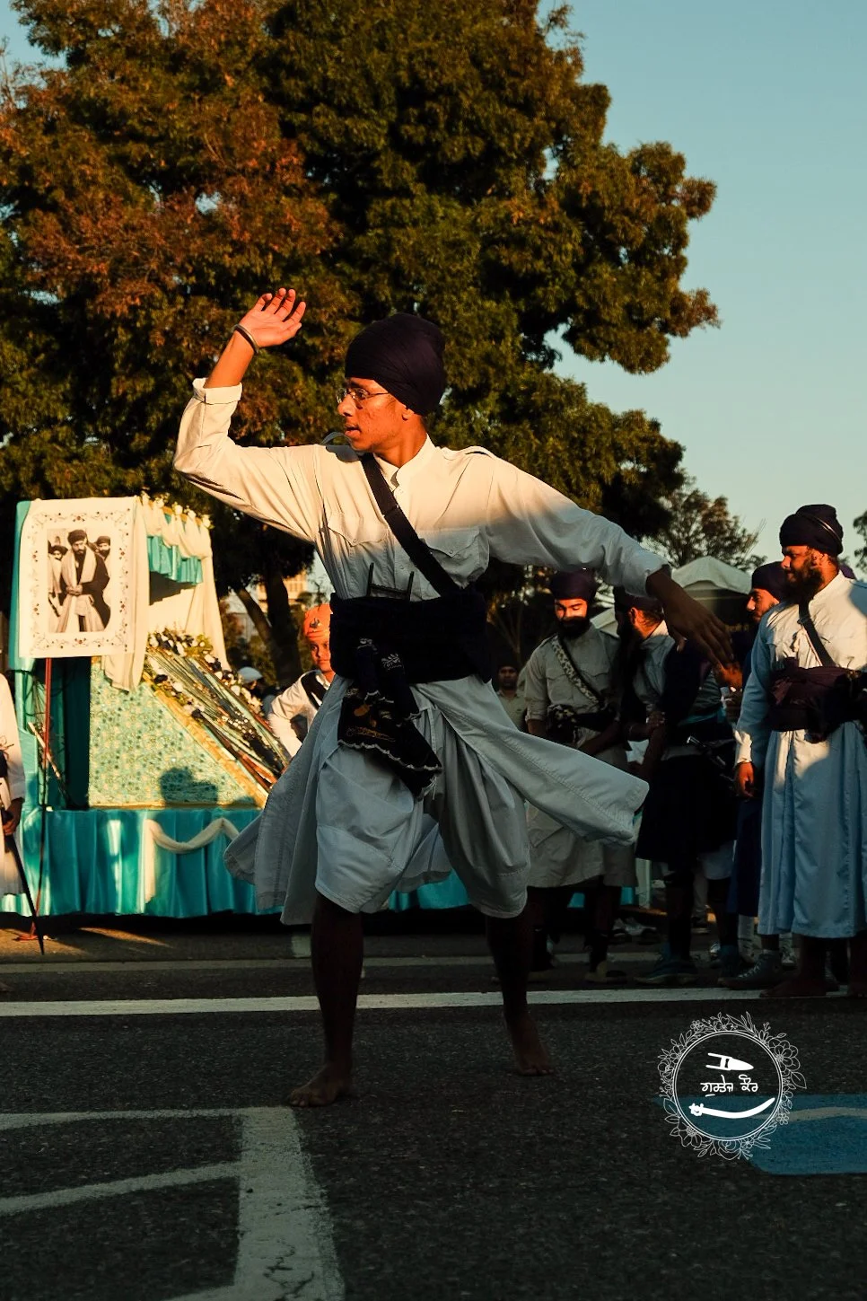 A man in traditional Sikh attire performing a martial arts move during a cultural event, with several other men in similar outfits watching. There are trees and a decorated display with a photo in the background.