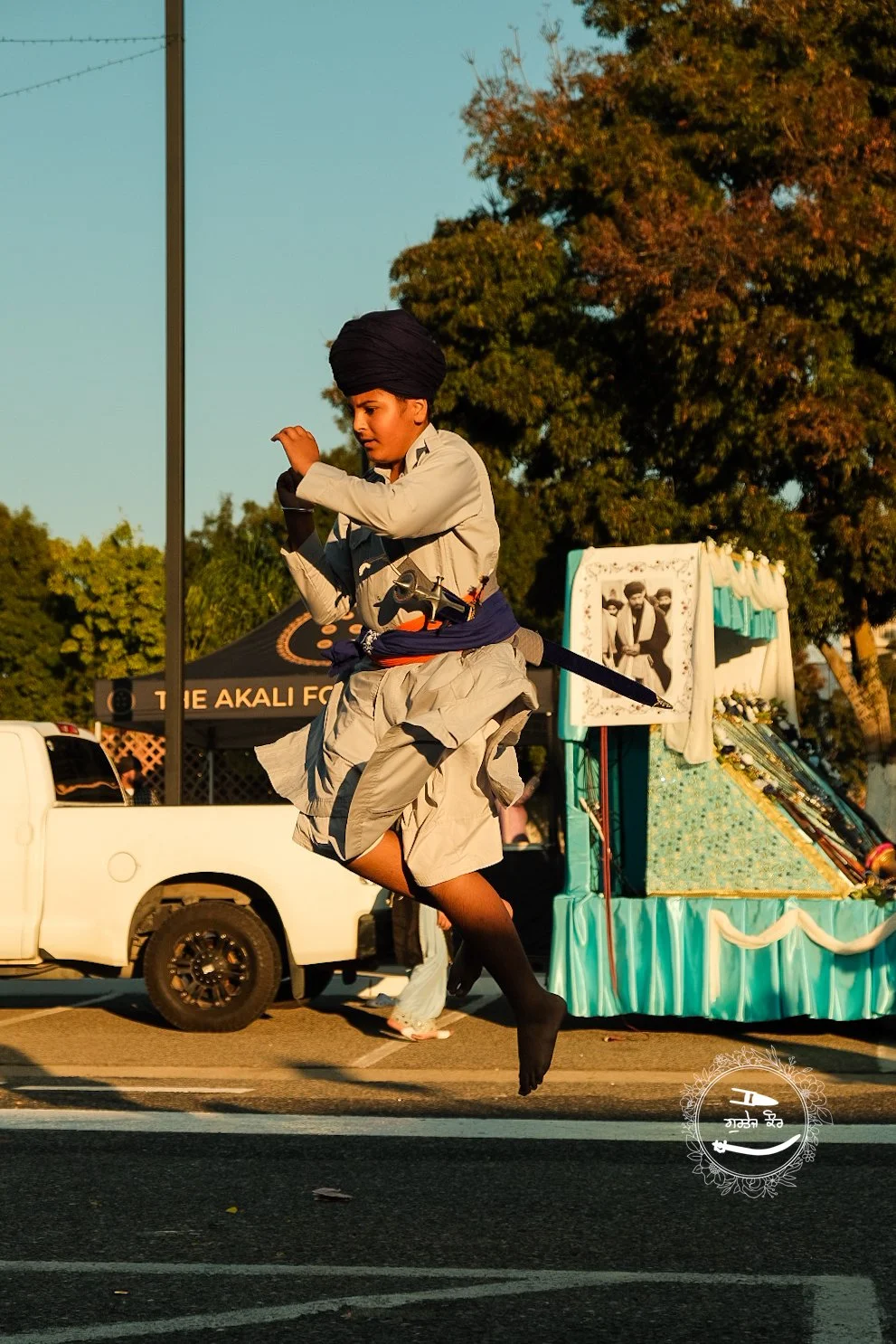 A man dressed in traditional Sikh attire, including a turban, is mid-air in a jump in front of a parade float decorated with pictures and fabric, during a street event.