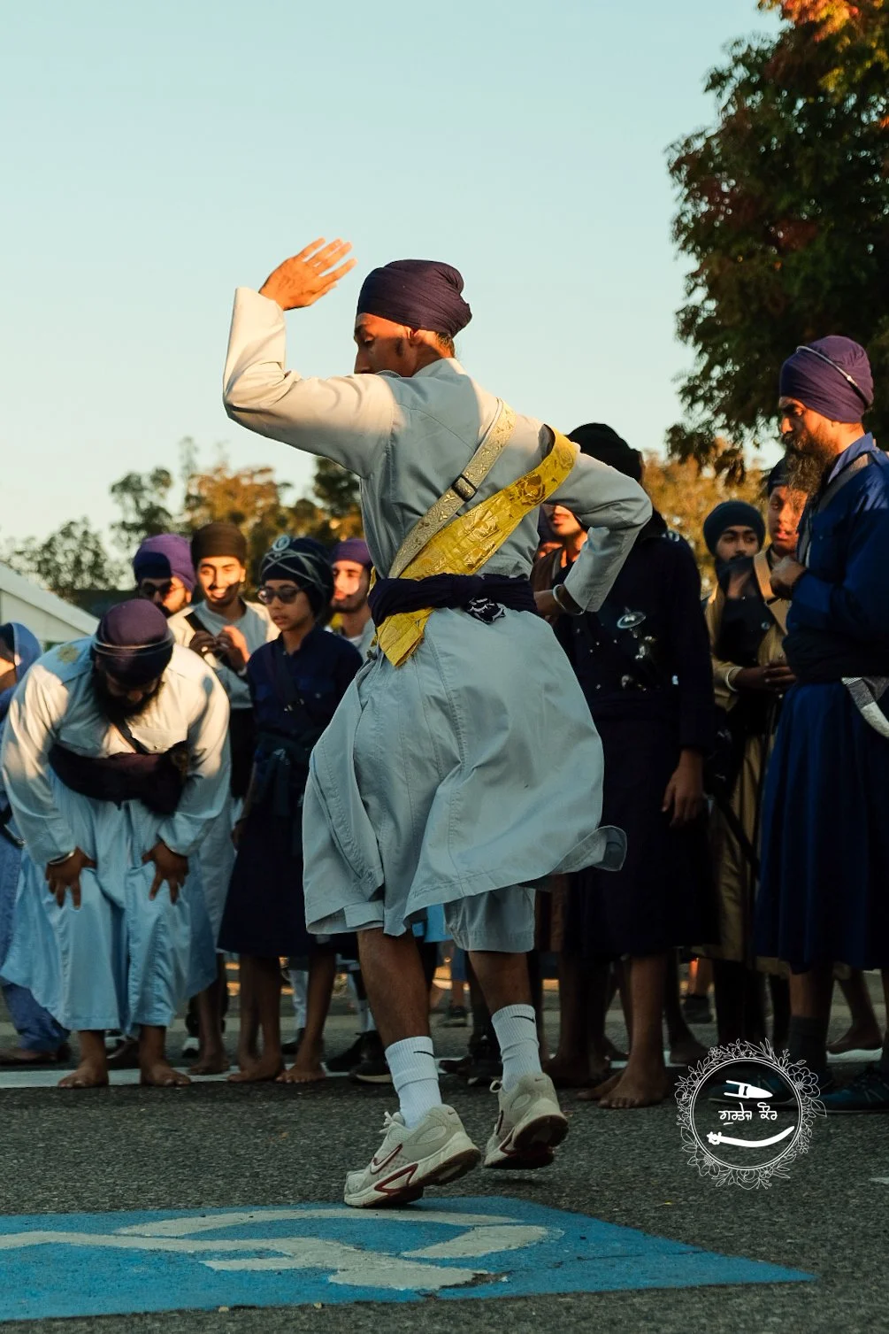 A man dressed in traditional Indian attire, wearing a turban, is dancing in front of a group of people also wearing traditional clothing. The group appears to be observing his dance, and the setting is outdoors during daylight, with trees in the back