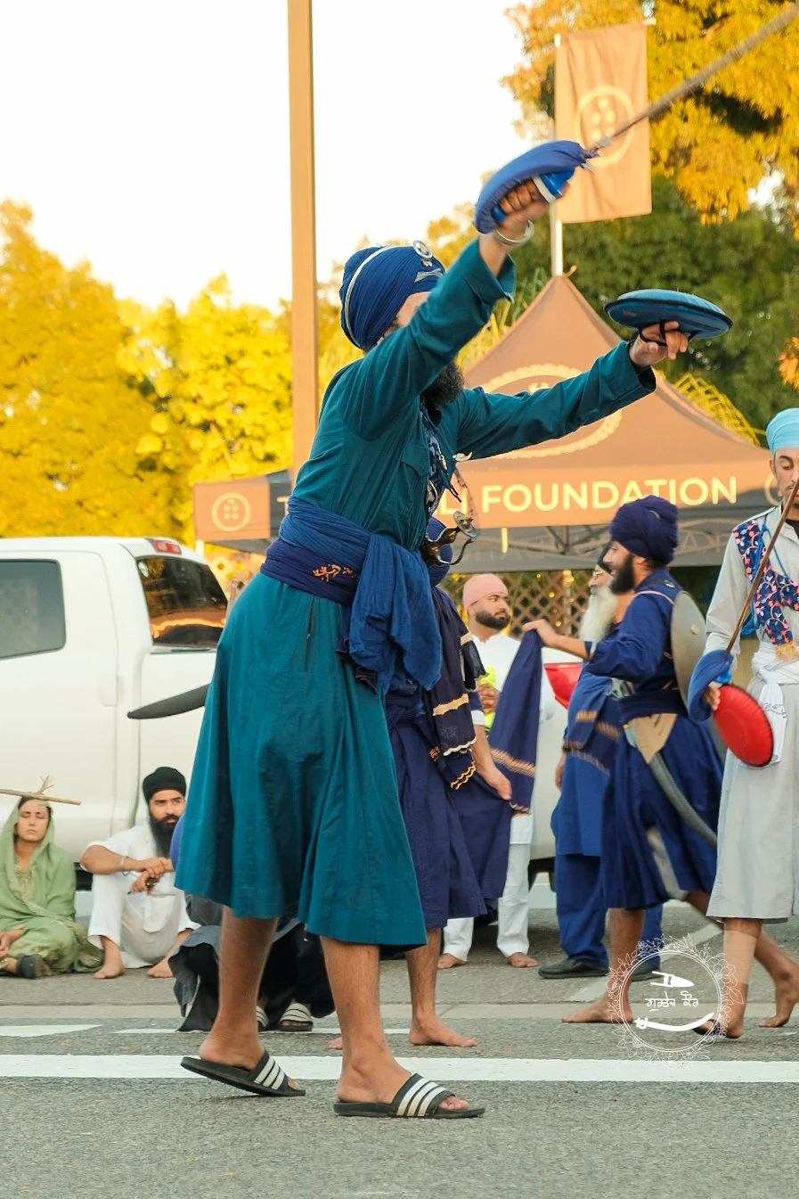 A group of people dressed in traditional Sikh attire participating in a street parade or celebration. Some are playing musical instruments, and the scene is set outdoors with trees showing fall foliage in the background.