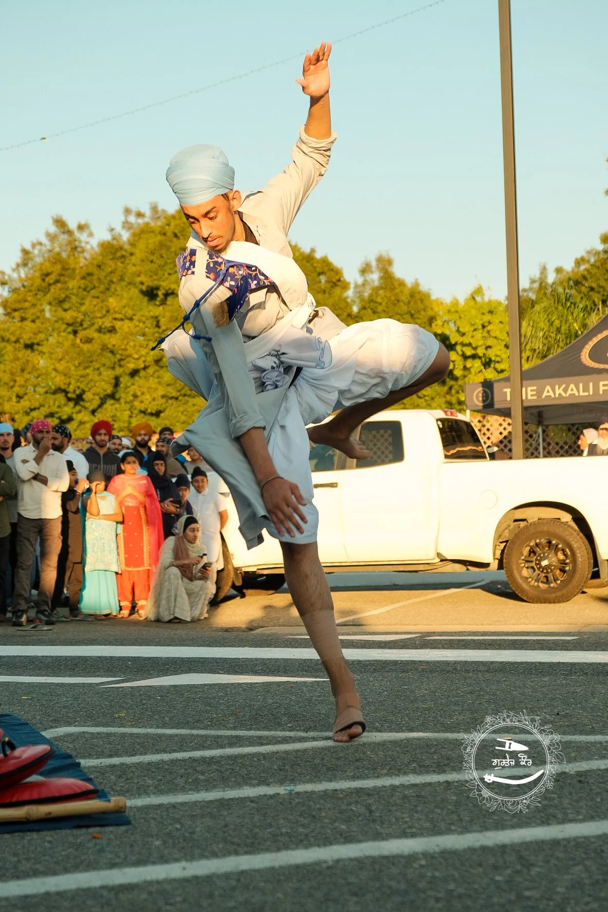 A man performing a martial arts move on a street, with a crowd of people watching behind him and a white truck parked nearby. The man is dressed in traditional attire and is balancing on one leg with an arm raised.