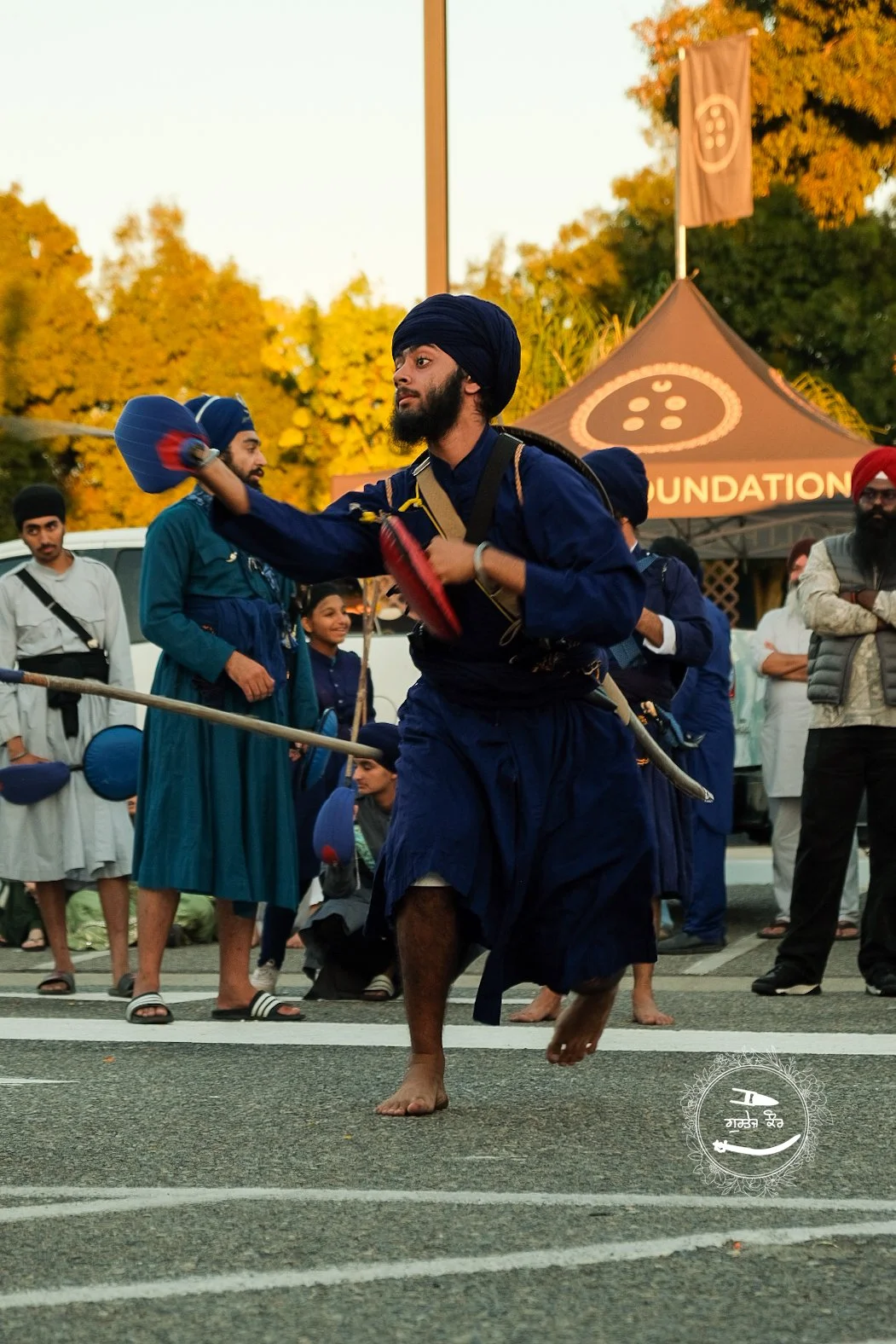 A man in traditional Sikh attire, wearing a blue turban and kurta, performing a martial arts display with a sword during a cultural event. Spectators watch in the background.