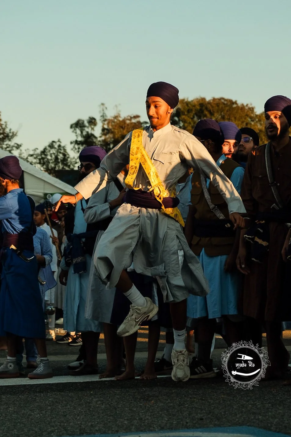 A young man in traditional Punjabi attire, including a turban and kurta, appears to be dancing or performing a jump at a cultural event with a group of people dressed in similar attire, outdoors during sunset.