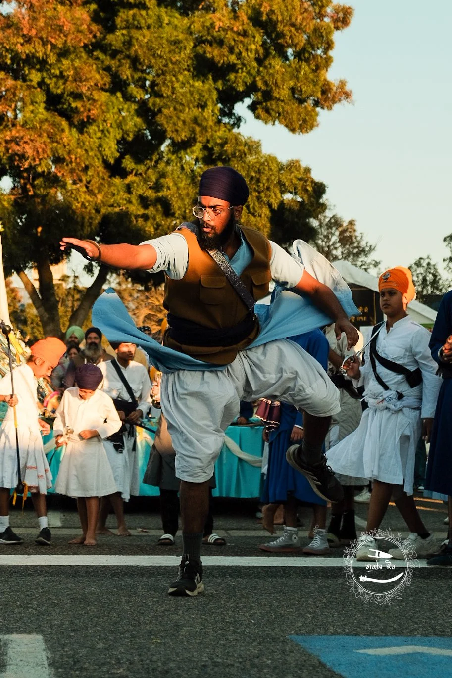 Man performing a traditional dance in Sikh attire at an outdoor cultural event, surrounded by onlookers in traditional clothing.