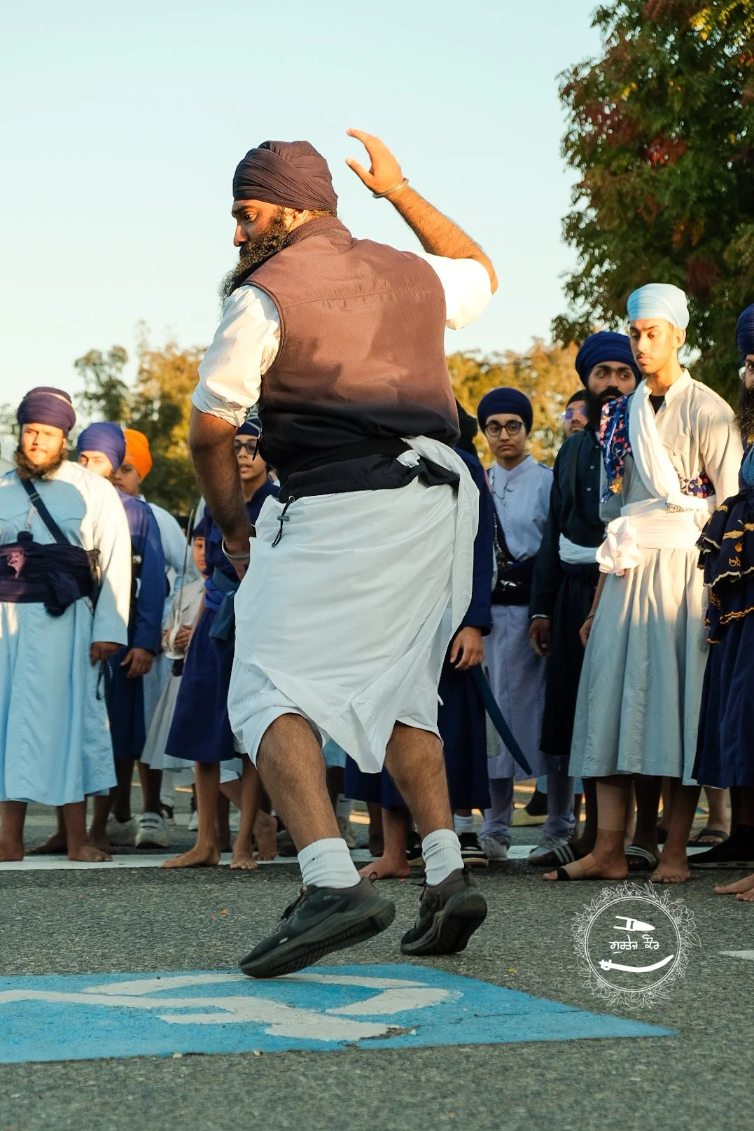 A man dancing on a blue wheelchair symbol on the ground in front of a group of people wearing traditional Sikh turbans and clothing, outdoors in daylight.
