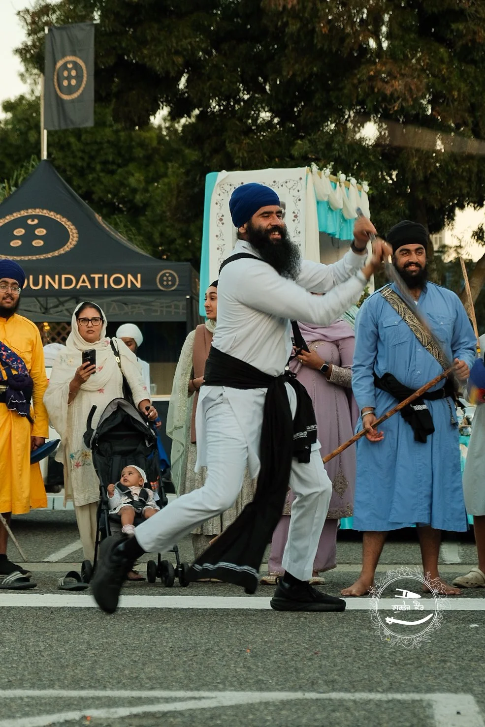 A group of people, some dressed in traditional Sikh clothing with turbans, are participating in a festival or parade outdoors. The scene includes a man smiling and swinging an object, with others standing around, some using their phones, and a baby s