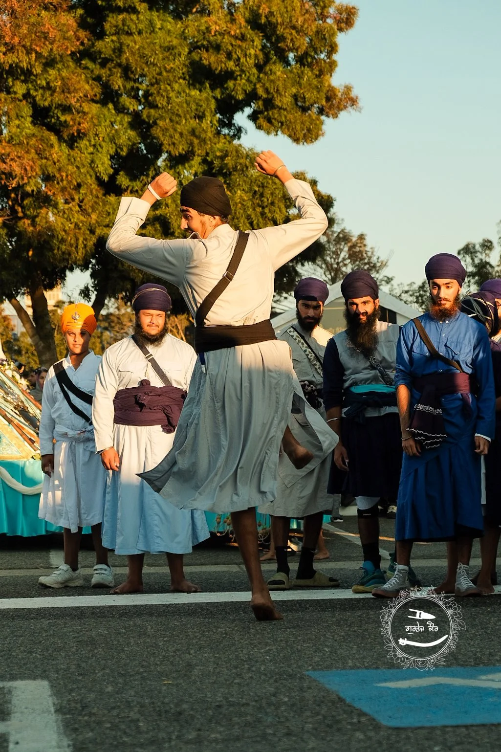 A man in traditional Sikh attire performs a dance or martial art move in front of a group of men, all wearing turbans, during an outdoor cultural event on a sunny day with trees in the background.