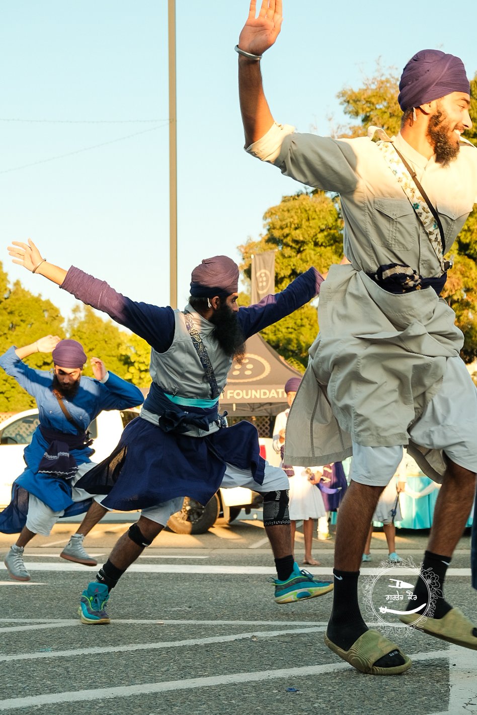 People participating in a traditional dance or performance on a street during daytime, wearing colorful costumes, with trees and a tent in the background.