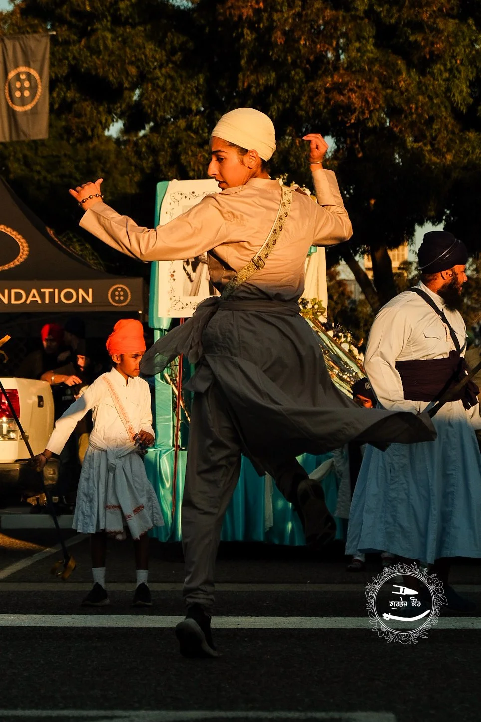 A woman in traditional Punjabi attire, dancing during a cultural event with others in similar attire in the background. There are trees and a tent with the word 'FOUNDATION' visible behind them.