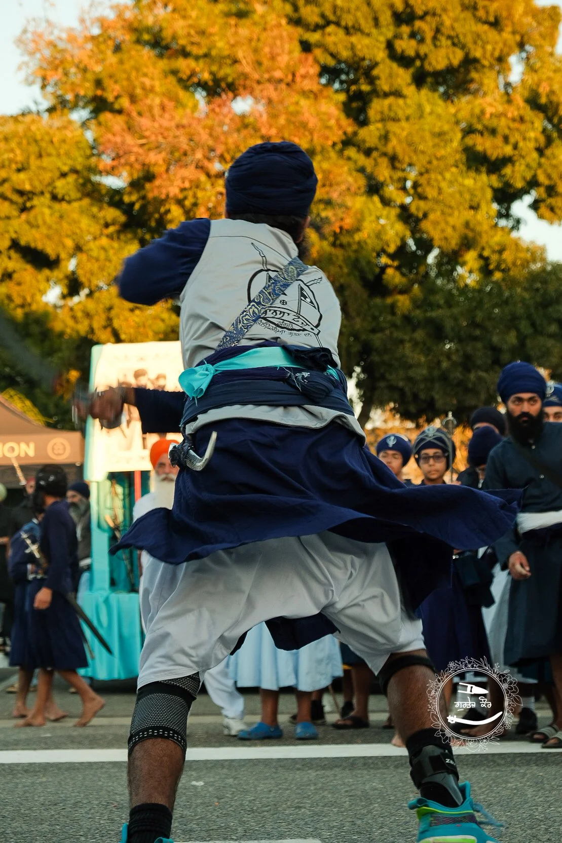 A man dressed in traditional Sikh attire engaging in a martial arts performance during an outdoor event, with onlookers in the background and a large tree with orange and yellow leaves overhead.