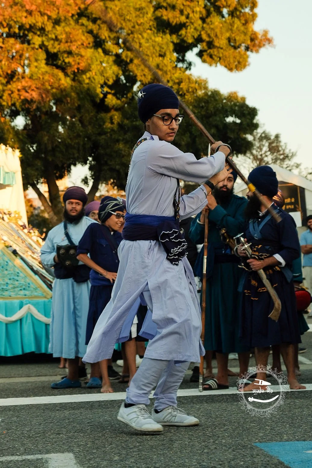 A group of people, most wearing traditional Sikh attire including turbans, standing on a street with a large tree and decorative setup in the background. One person in the foreground, wearing glasses and a turban, is performing a martial arts move wi