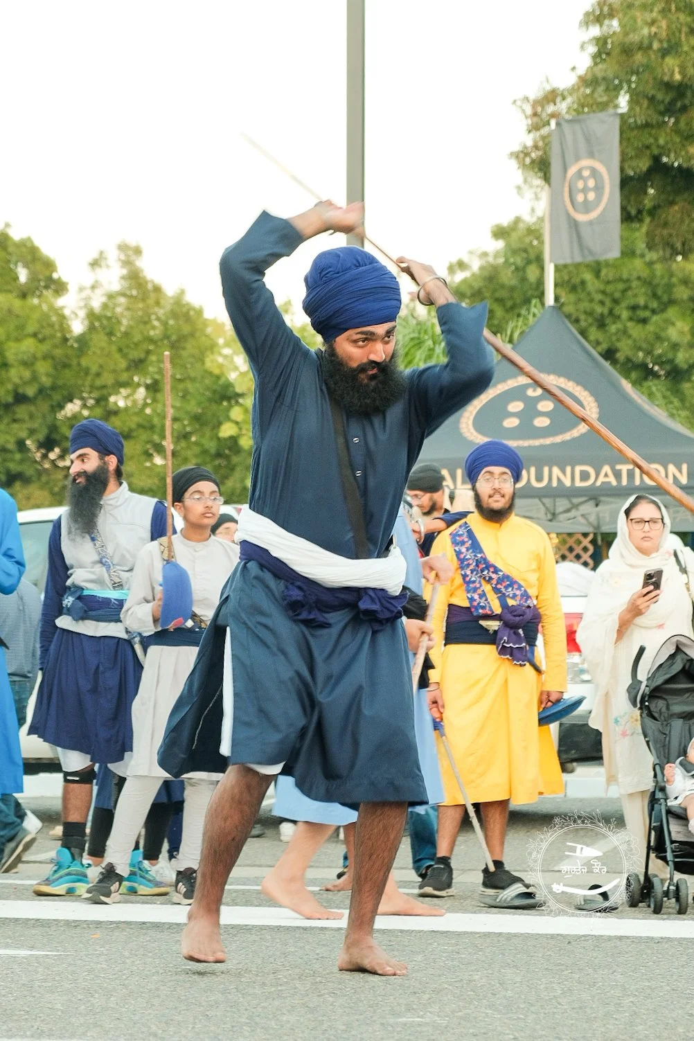 A man dressed in blue traditional Indian attire performing a dance with a long stick in a street parade. He is wearing a blue turban and has a beard. Several other people in colorful traditional clothing and turbans are watching him, some holding sti