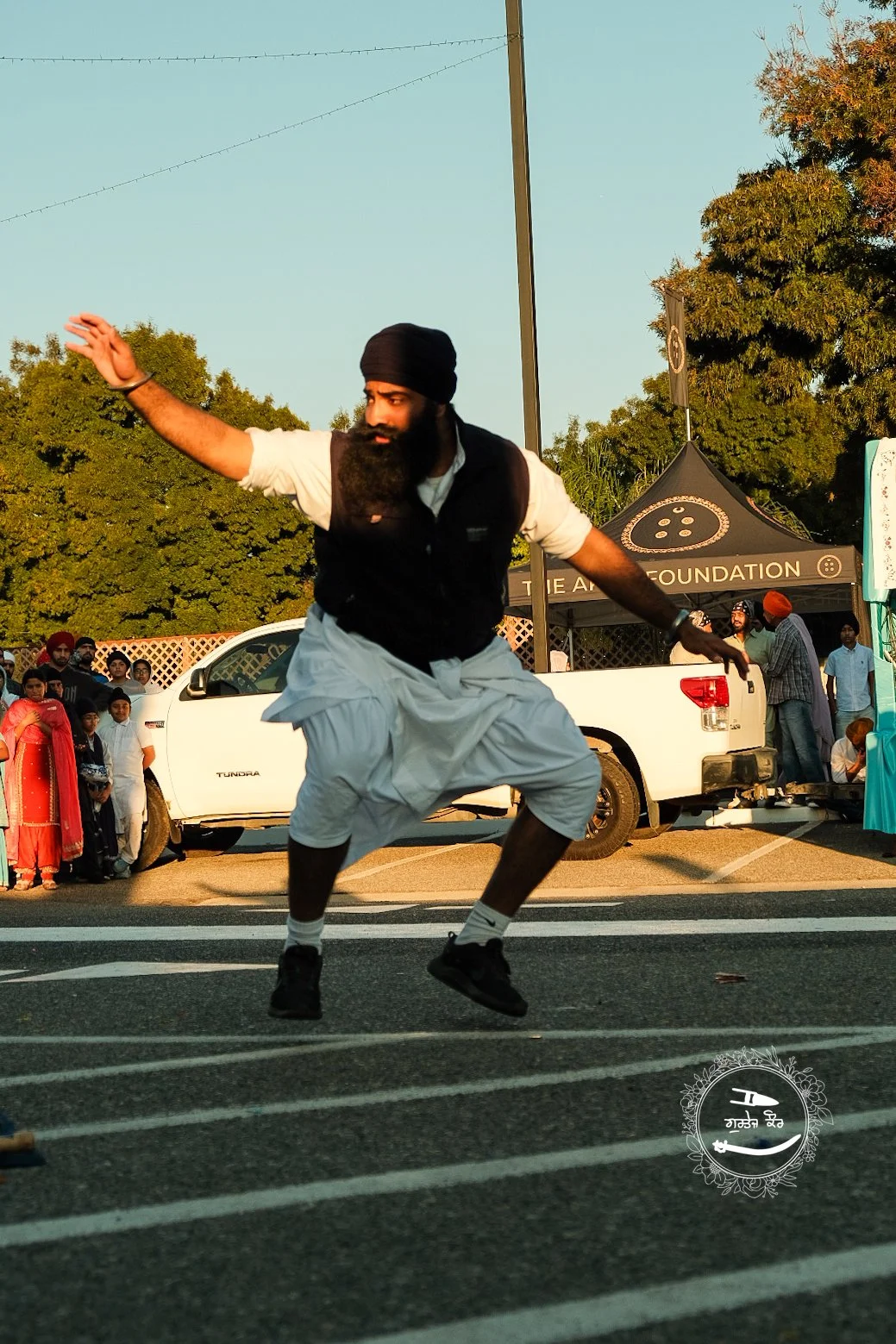A man in traditional Indian attire, including a turban, performing a dance or a jump on a street during an outdoor event, with onlookers and event tents in the background.