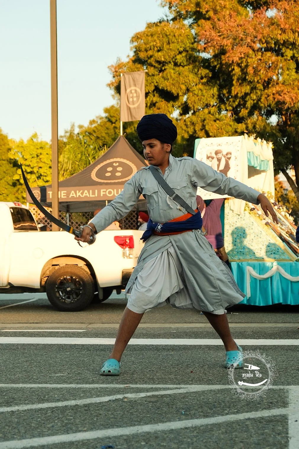 Boy dressed in traditional Sikh attire performing Gatka with a sword during a parade or festival, with tents and trees in the background.