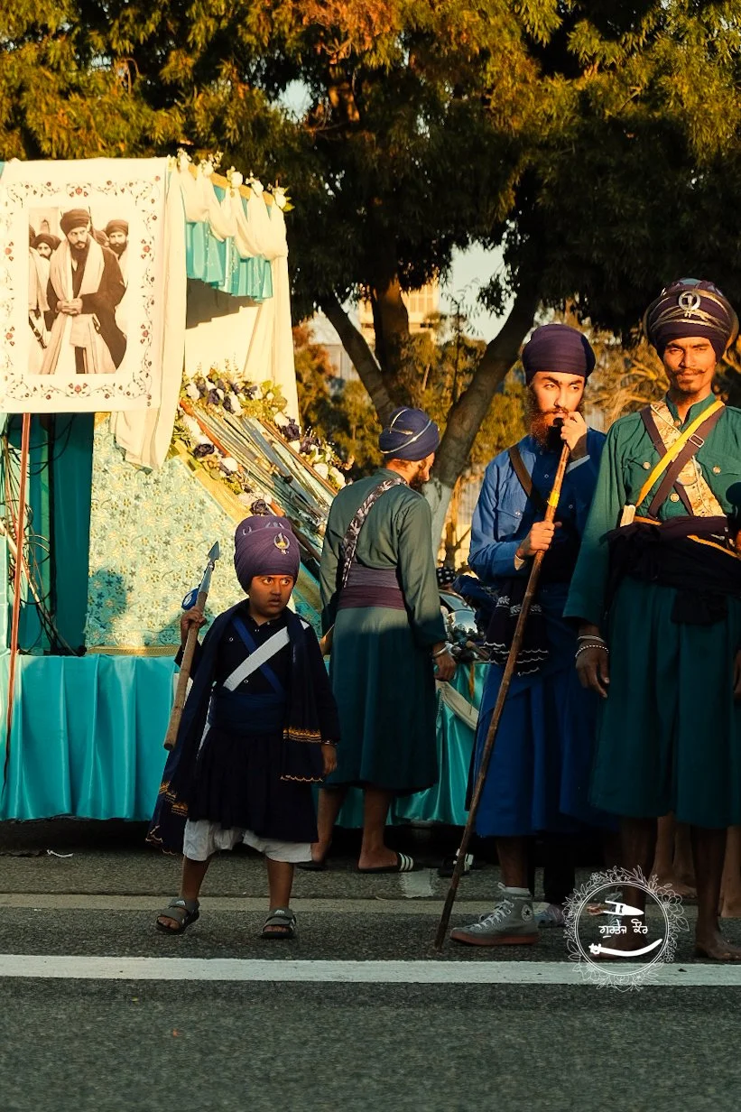 Group of men and child in traditional Sikh attire participating in a parade or procession, with a decorated float displaying religious or cultural images and items, set outdoors during daylight.