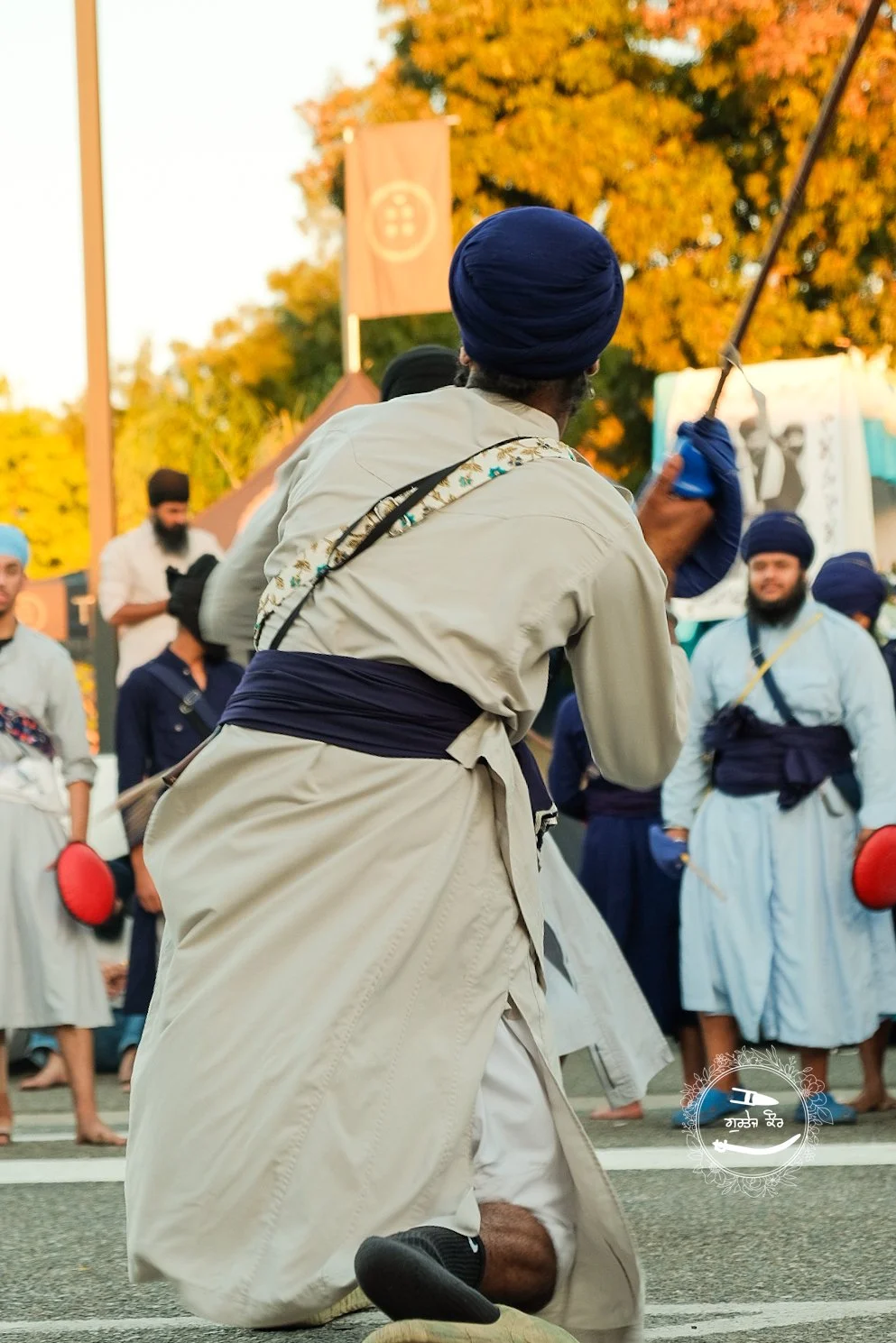 A man kneeling on the ground, wearing a turban and traditional clothing, holding a stick, with a group of men in turbans and traditional attire in the background during an outdoor event in autumn.