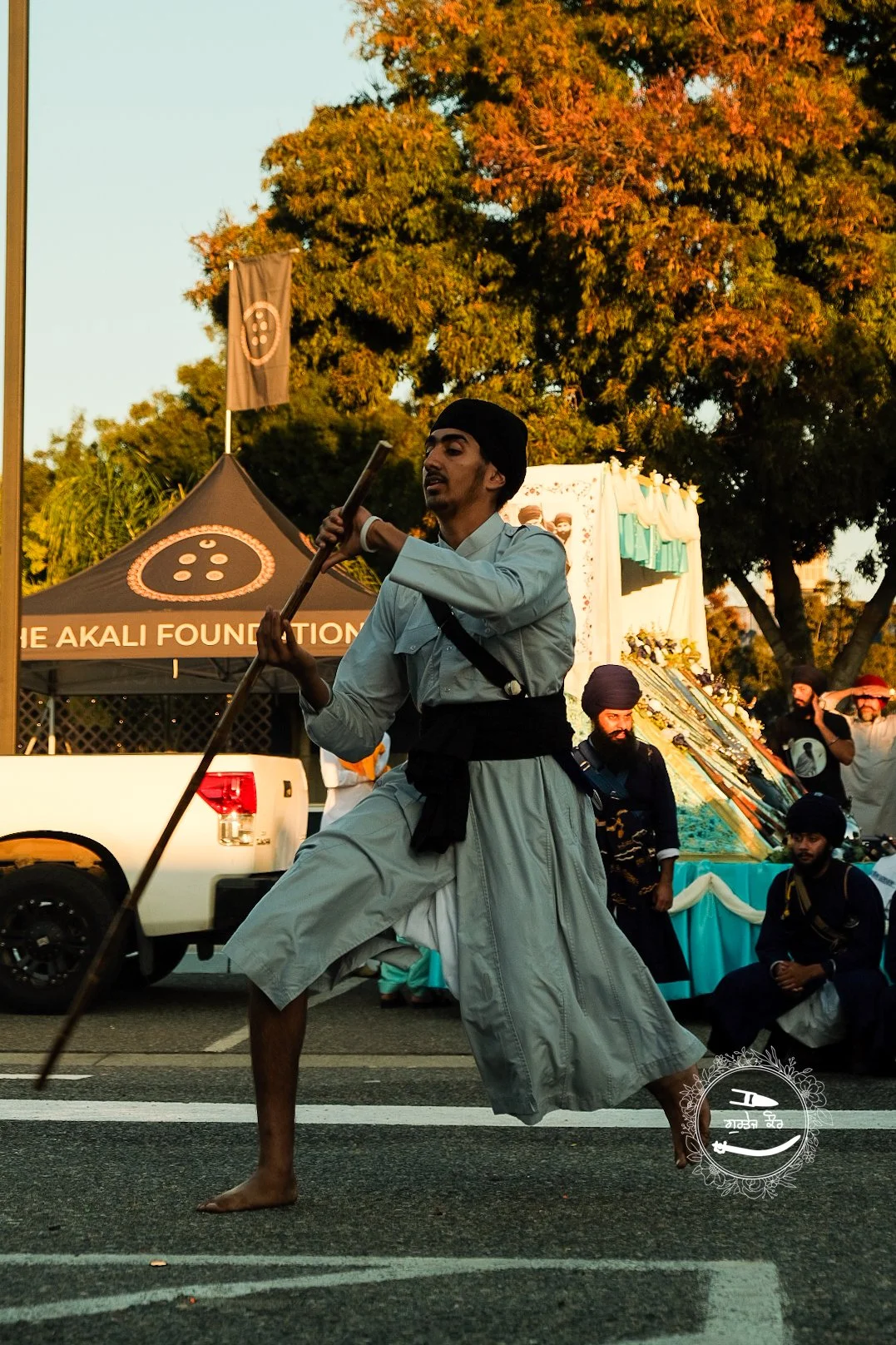 A man performing a traditional dance in the street during a celebration, with a backdrop of trees, a tent, and people watching.
