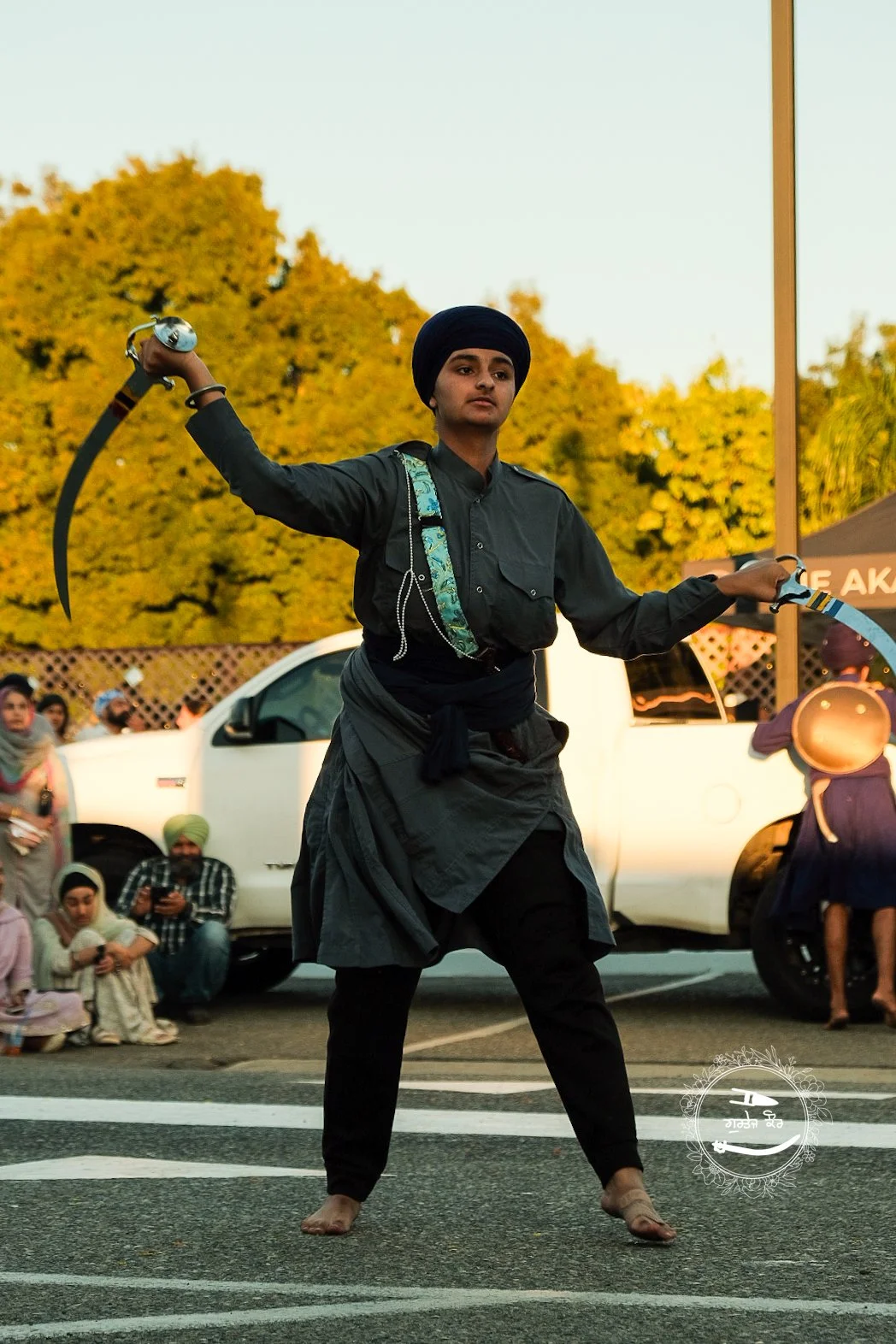 A person dressed in traditional black attire performing a street dance with two curved blades, standing on a crosswalk while onlookers watch in the background.
