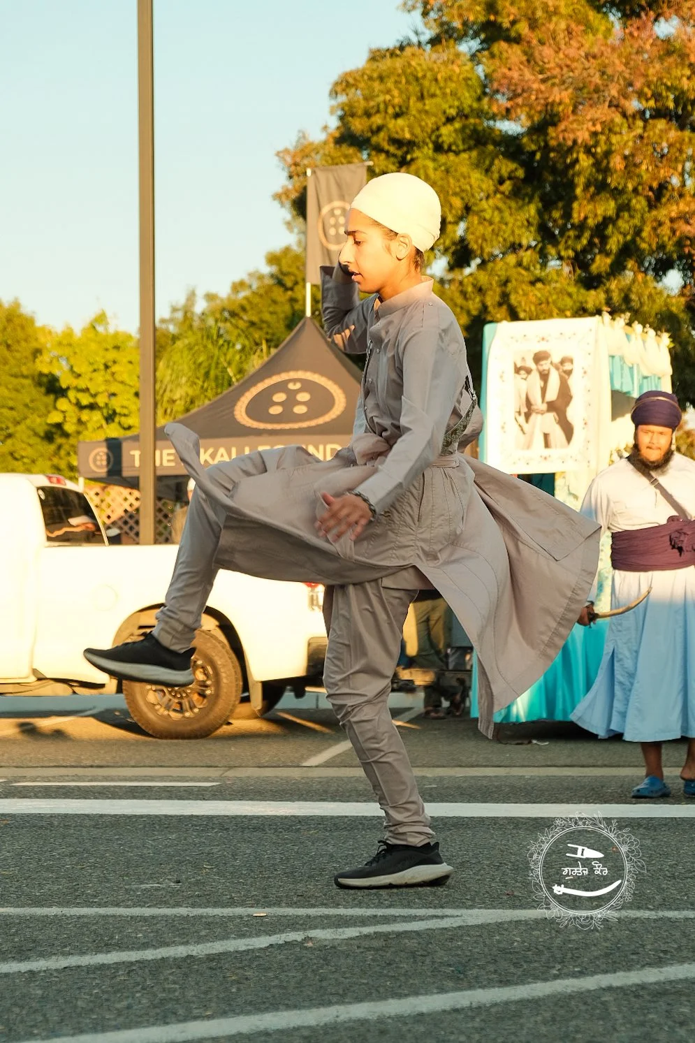 A person dressed in traditional attire, wearing a white turban, is performing a dance move on a street during a cultural event. The scene includes a tent in the background, trees, and another person dressed in traditional clothing.