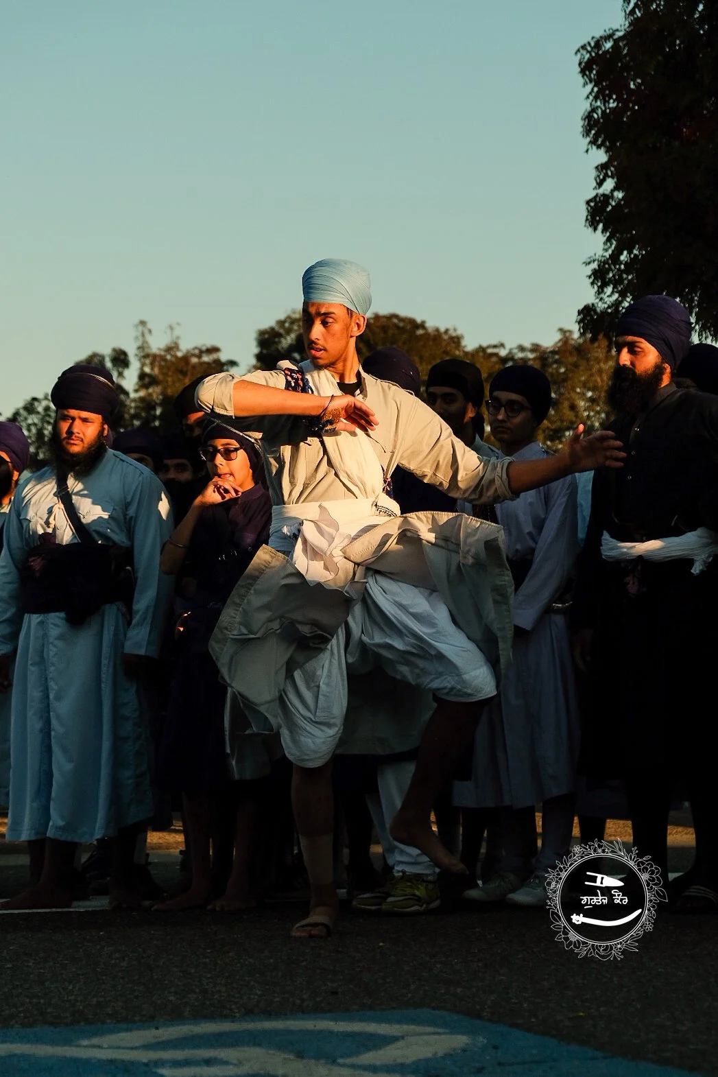 A group of Sikh men and women gathered outdoors during sunset, with a man in the center performing a traditional martial arts move, kicking in the air while others watch.