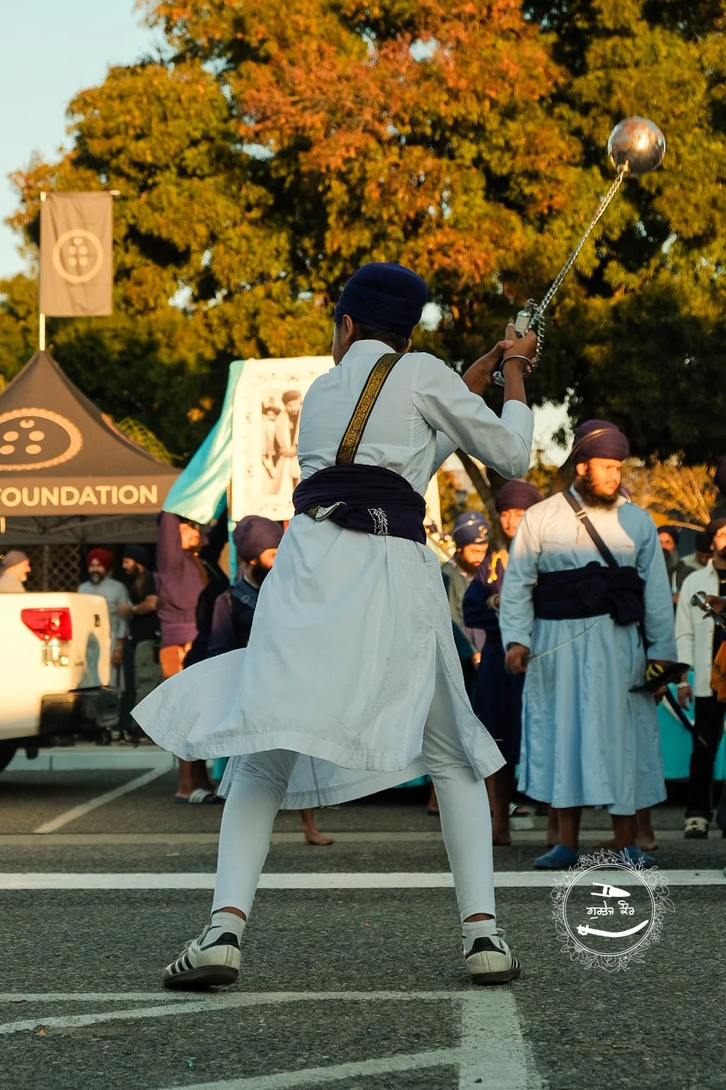 A person dressed in traditional Sikh attire, including a white kurta, white pants, and a blue turban, performs a martial arts or dance move with a metal chain weapon during a cultural event. There are several other people in similar attire in the bac