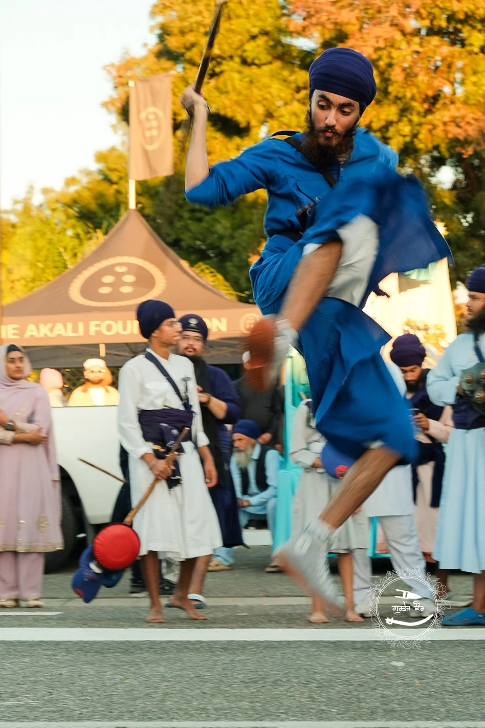 A man in blue traditional attire and a blue turban is leaping in the air during a cultural event, with people in traditional dresses and a tent with the logo of 'The Akali Foundation' in the background.