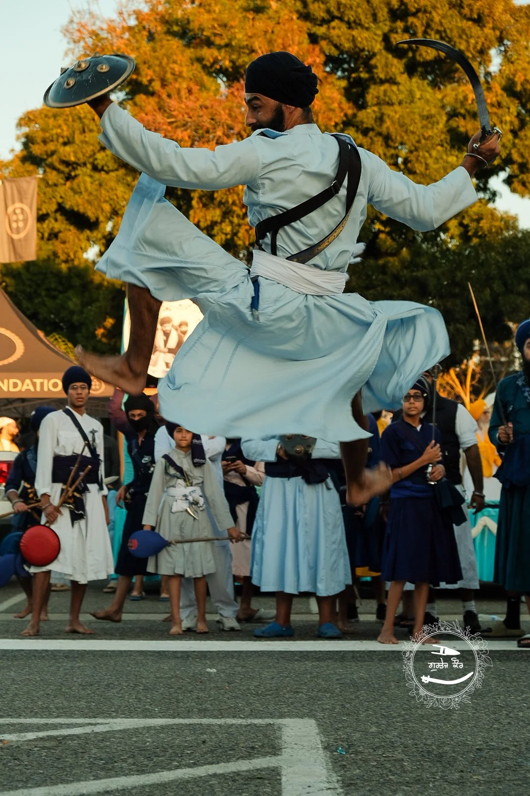 A man wearing traditional Sikh white attire, with a turban, is jumping in the air performing a martial arts or cultural dance in front of a crowd, with autumn trees in the background.
