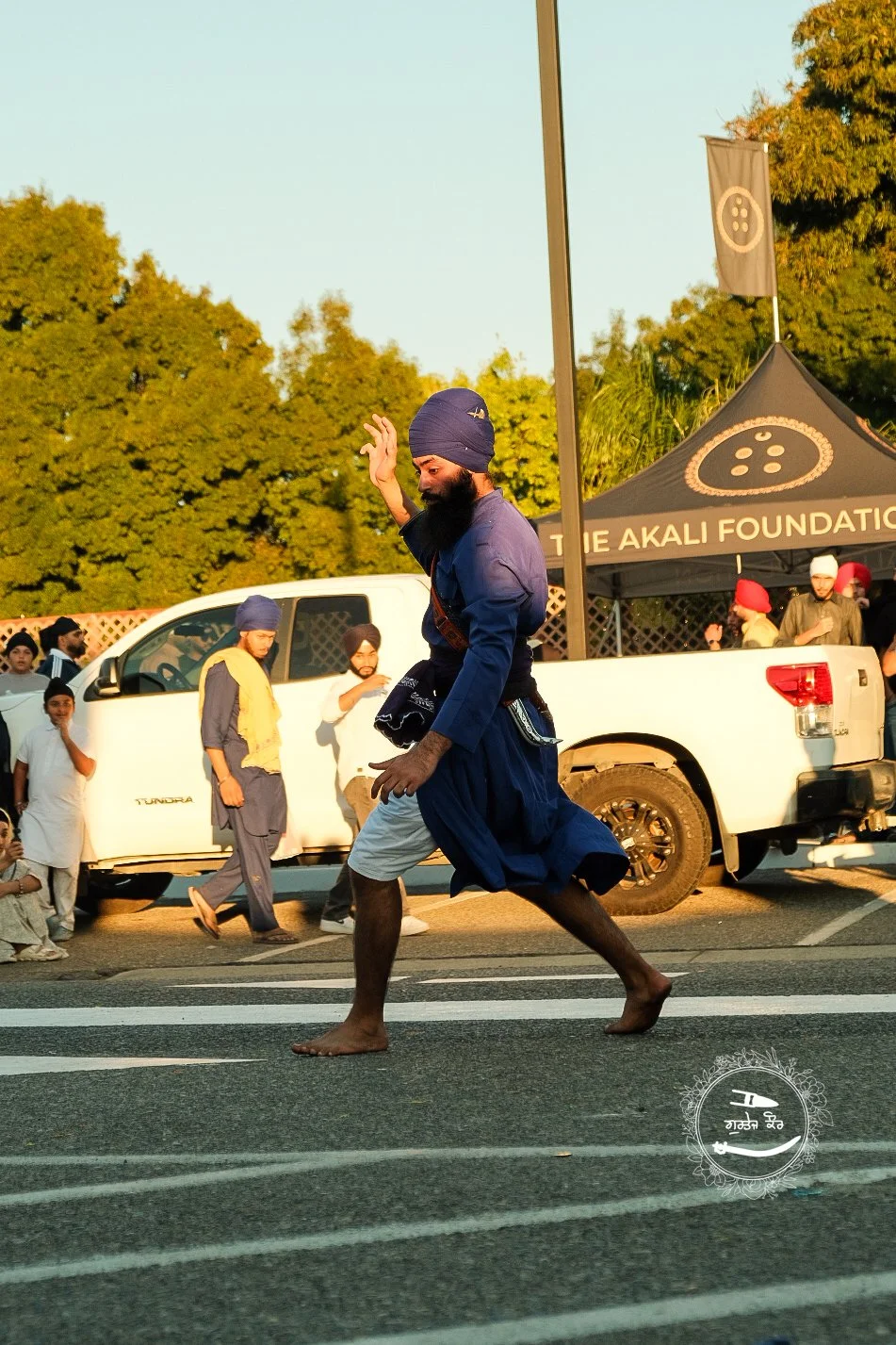 A man wearing a blue turban and traditional blue and white clothing performing a dance on the street, with a crowd and a white pickup truck in the background, exhibiting a cultural event or festival.