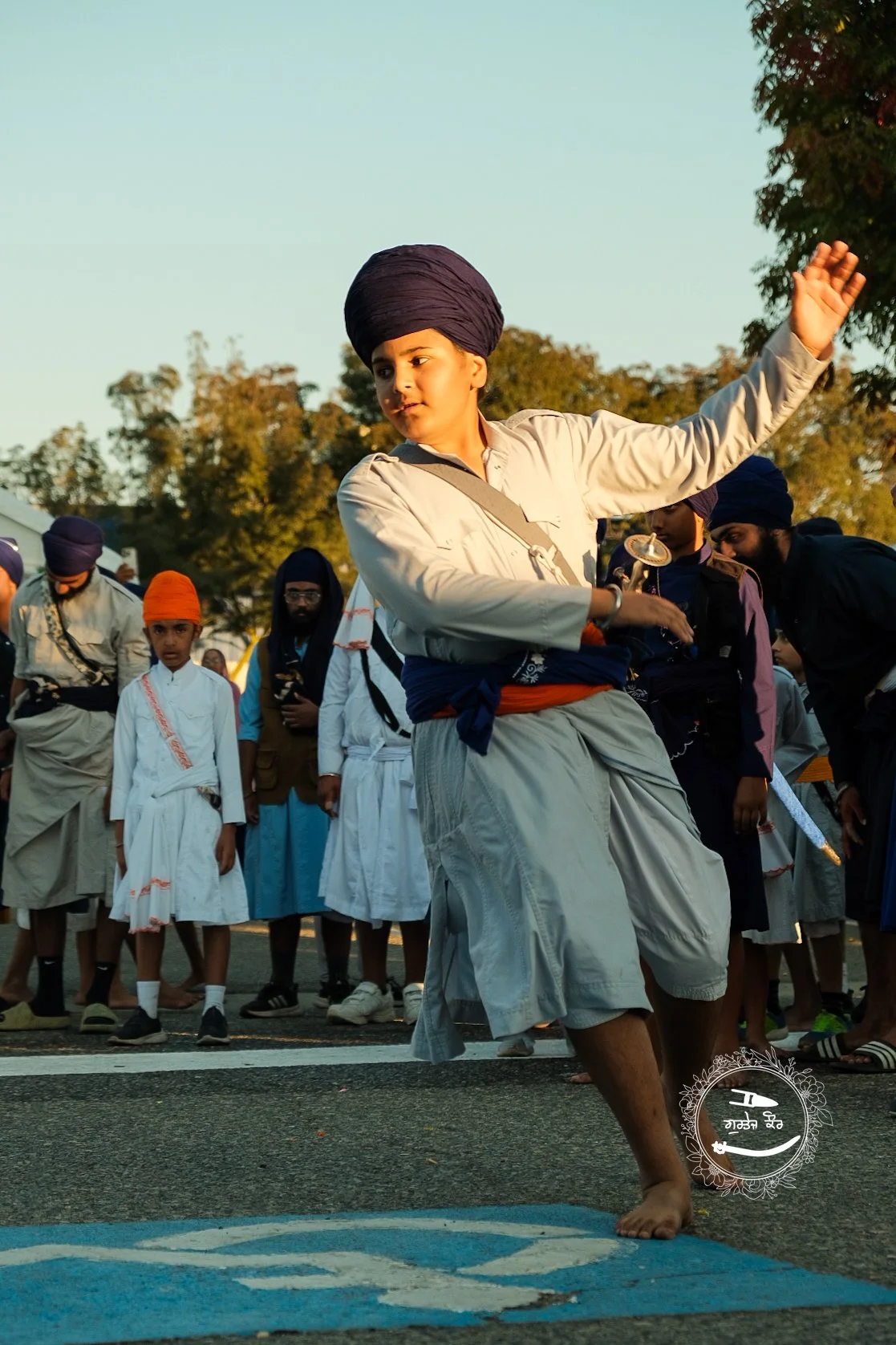 Young boy in traditional Sikh attire performing a dance in a parade, surrounded by onlookers dressed in similar traditional clothing, with trees and a clear sky in the background.