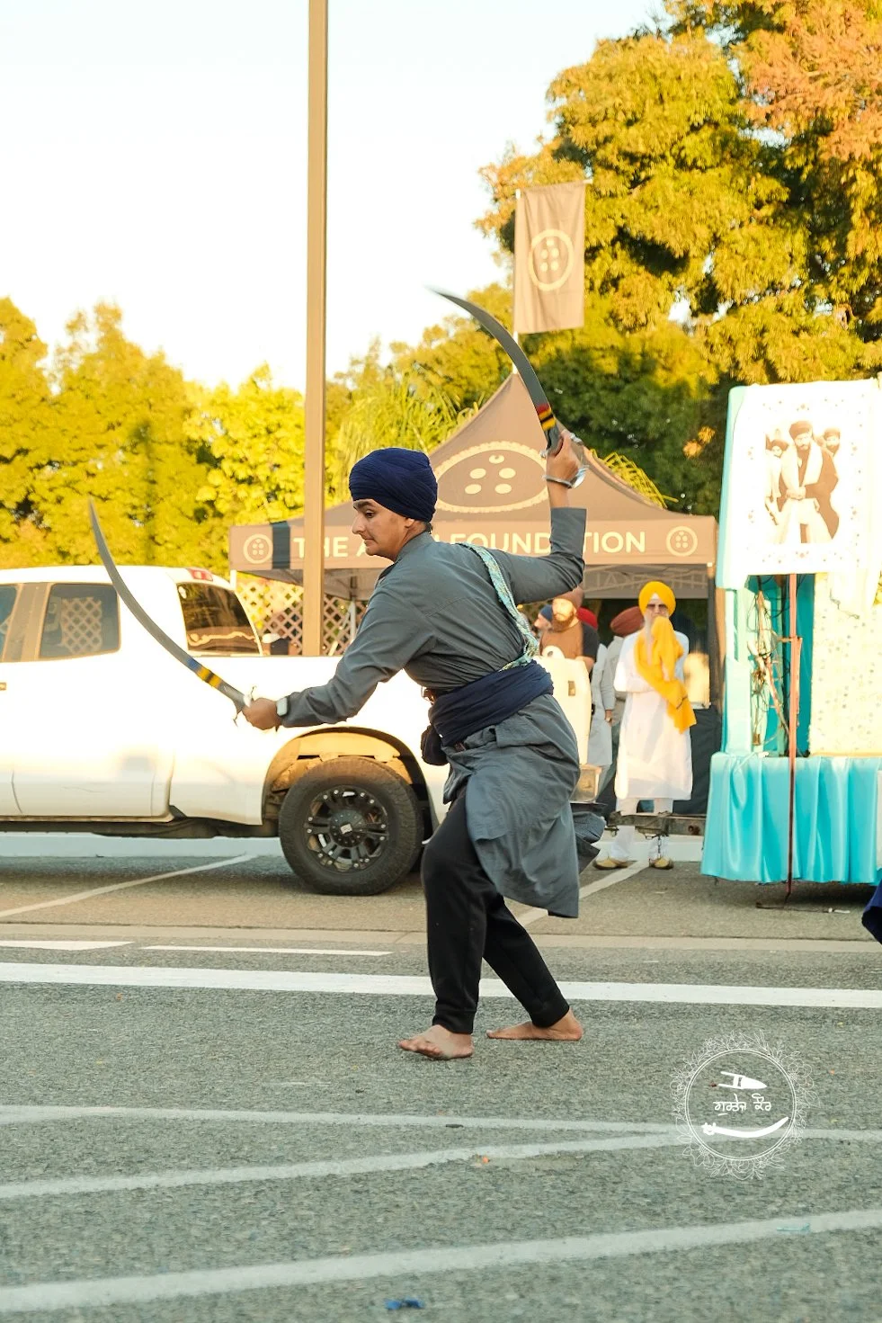 A man dressed in a dark turban and traditional Sikh attire performing a martial arts move with swords on a street, with a group of people in the background near a tent.