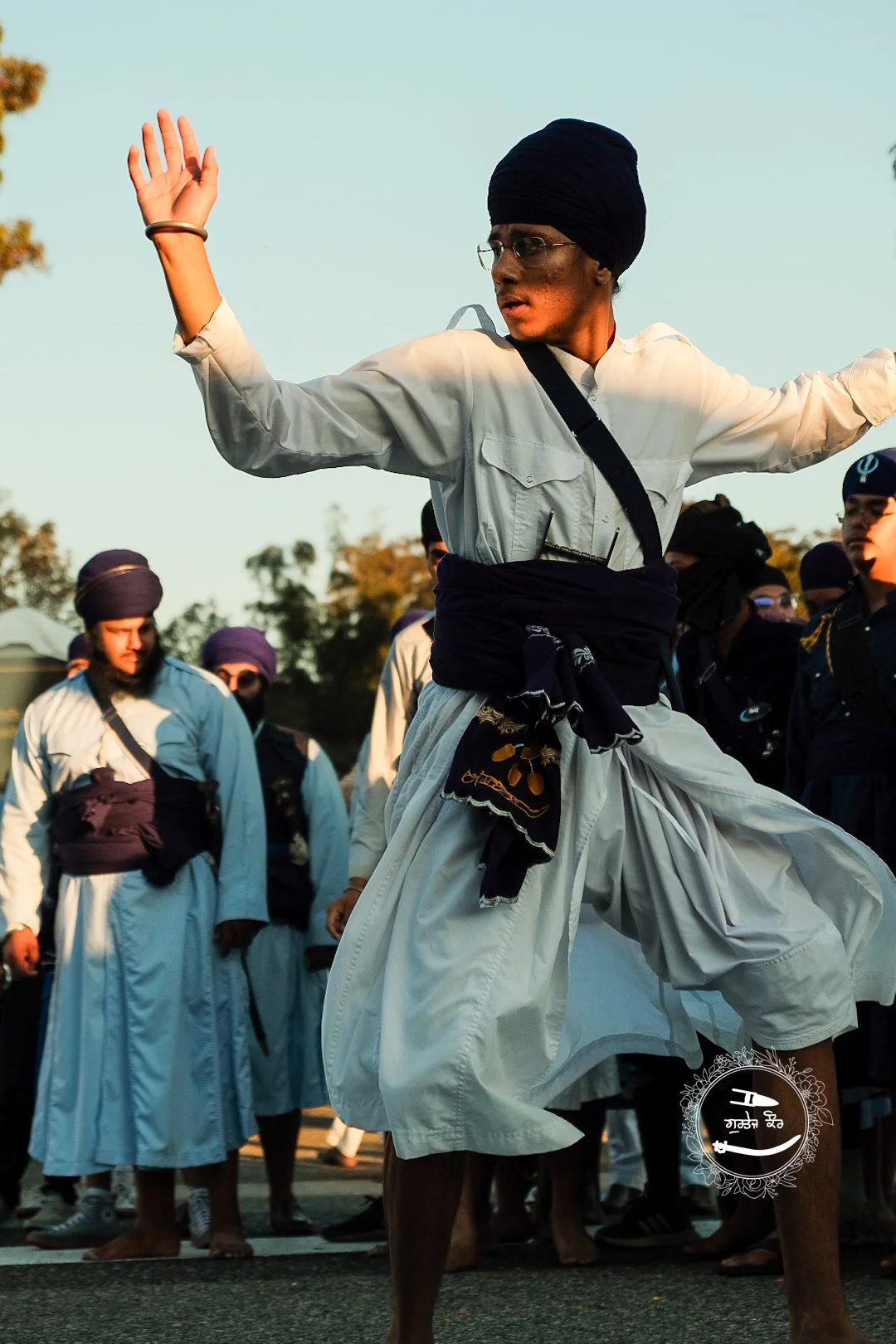 Man wearing a turban and traditional white attire performing a dance at an outdoor gathering with other people dressed similarly in the background.