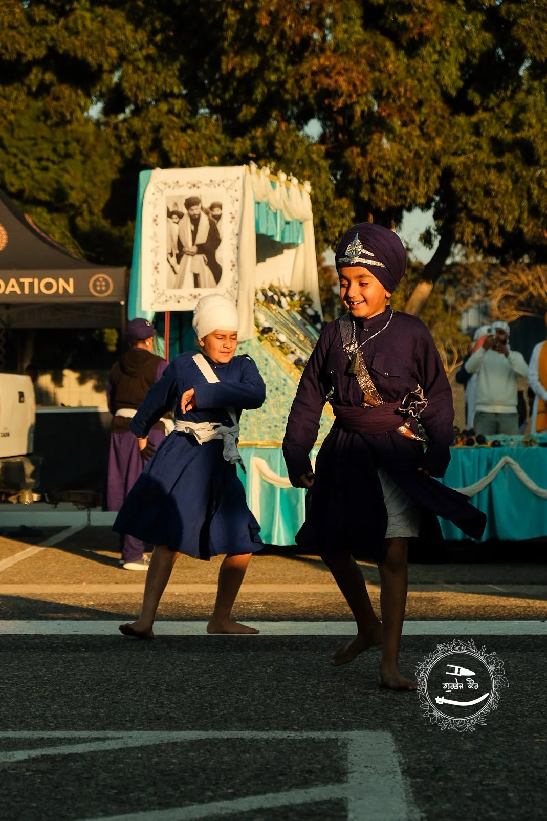 Two children dressed in traditional Sikh attire, dancing barefoot on the street during a celebration, with a decorated float and crowds in the background.