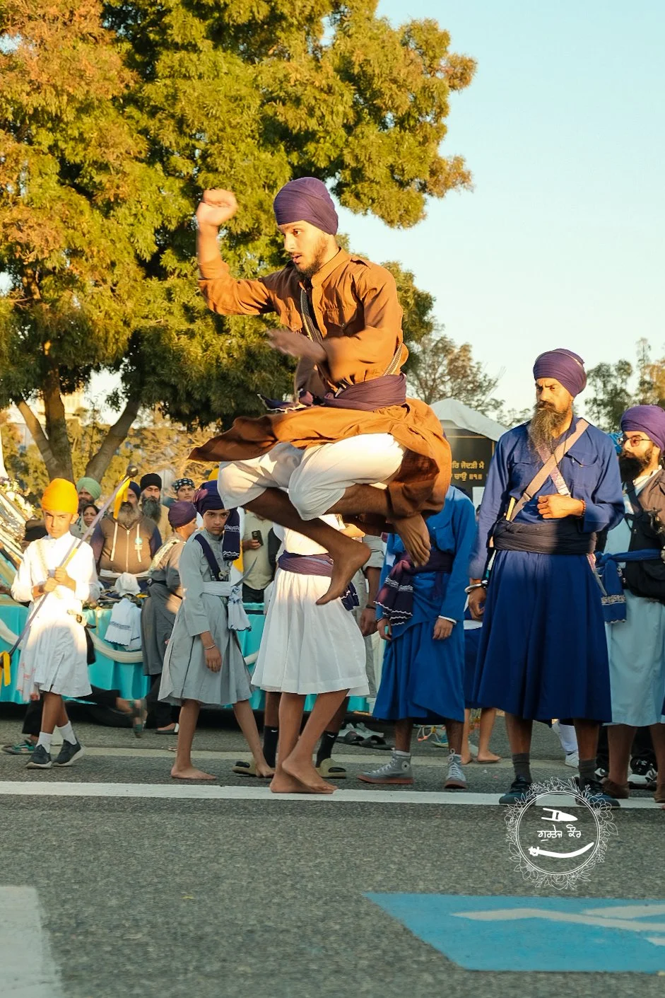 A man dressed in traditional Sikh attire is captured mid-air in a jump during a parade or gathering, with others in similar attire and turbans watching and participating in the event, set outdoors with trees and a clear sky.