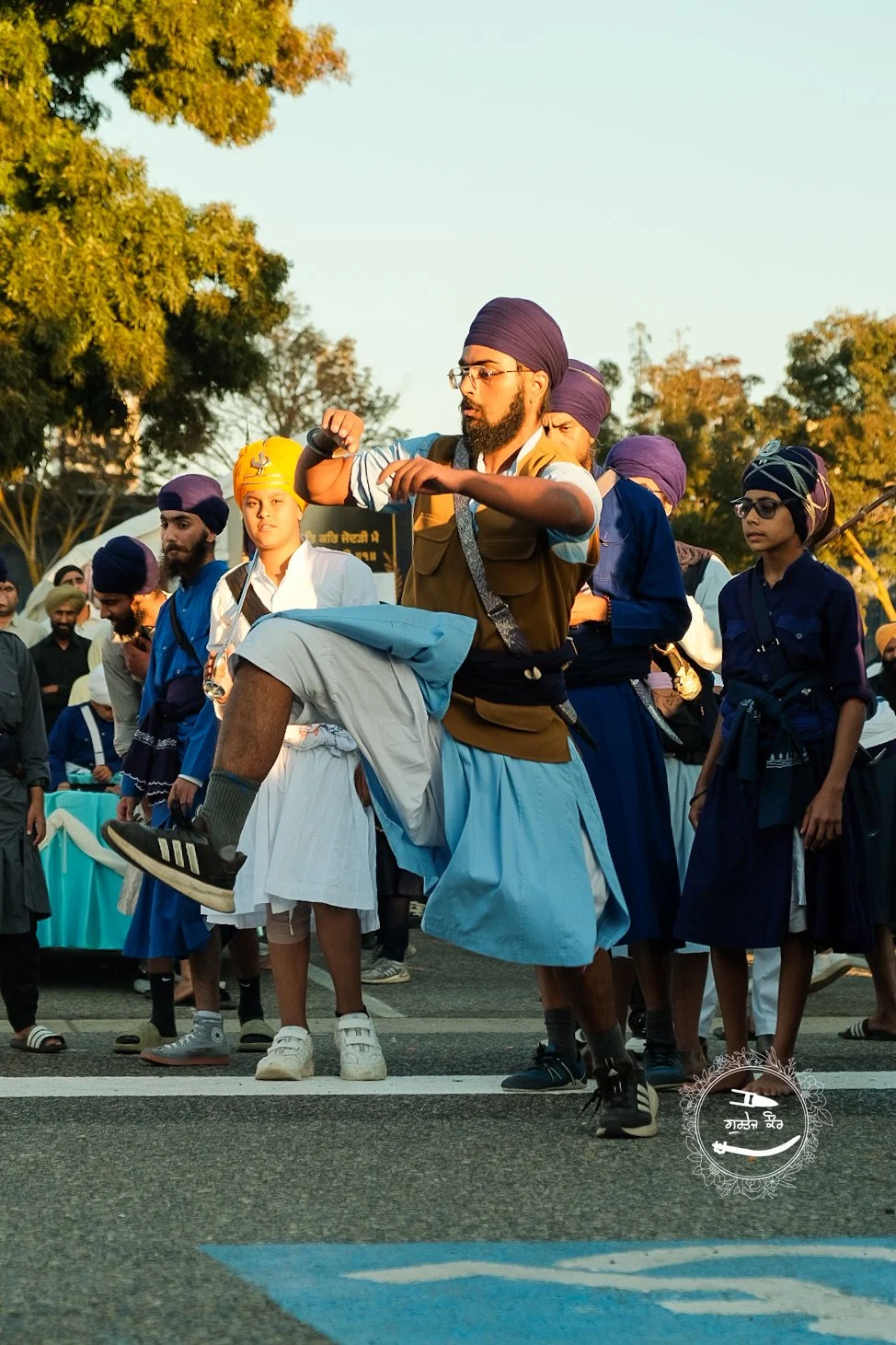 A group of men and boys wearing traditional Sikh turbans standing on a street during daytime, with one man in the foreground performing a dance move.