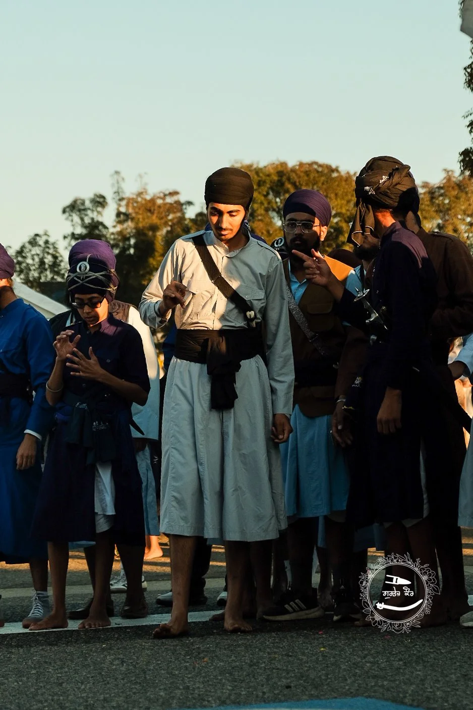 Group of young men and boys dressed in traditional Sikh attire standing outdoors, some looking down or engaged in conversation, with trees and a clear sky in the background.