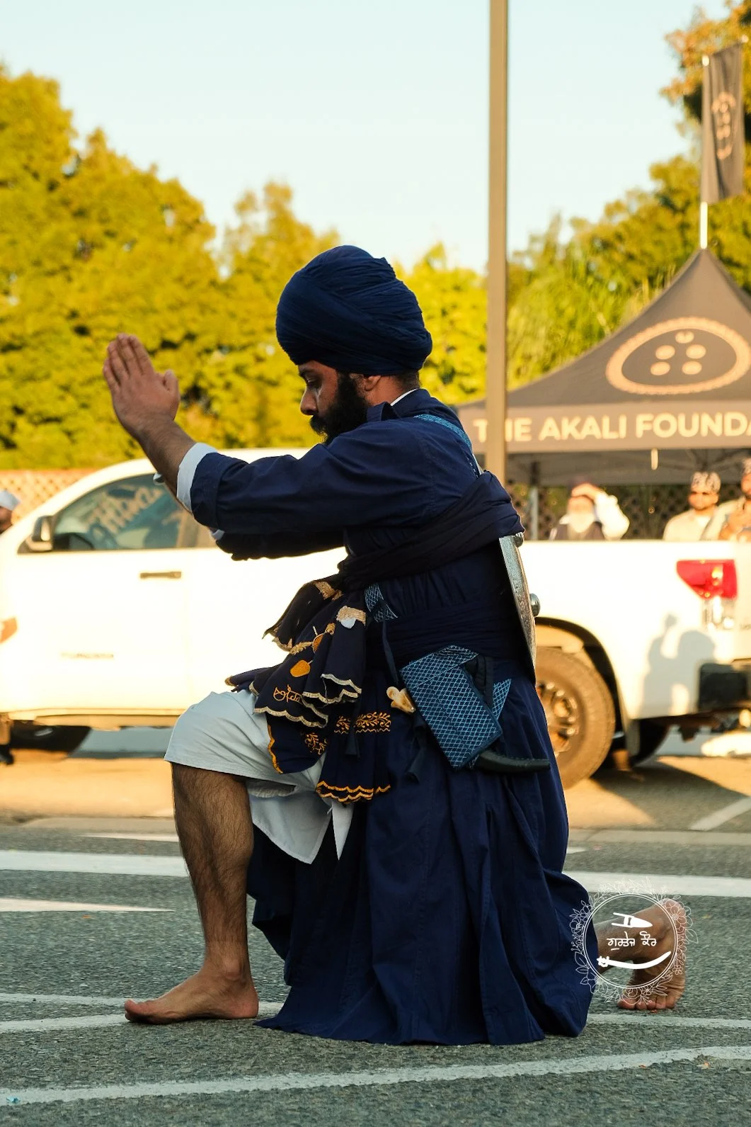 A man dressed in traditional Indian attire, including a blue turban and clothing, kneeling on a street and performing a prayer or ritual with his hands raised. There are cars and a tent in the background.