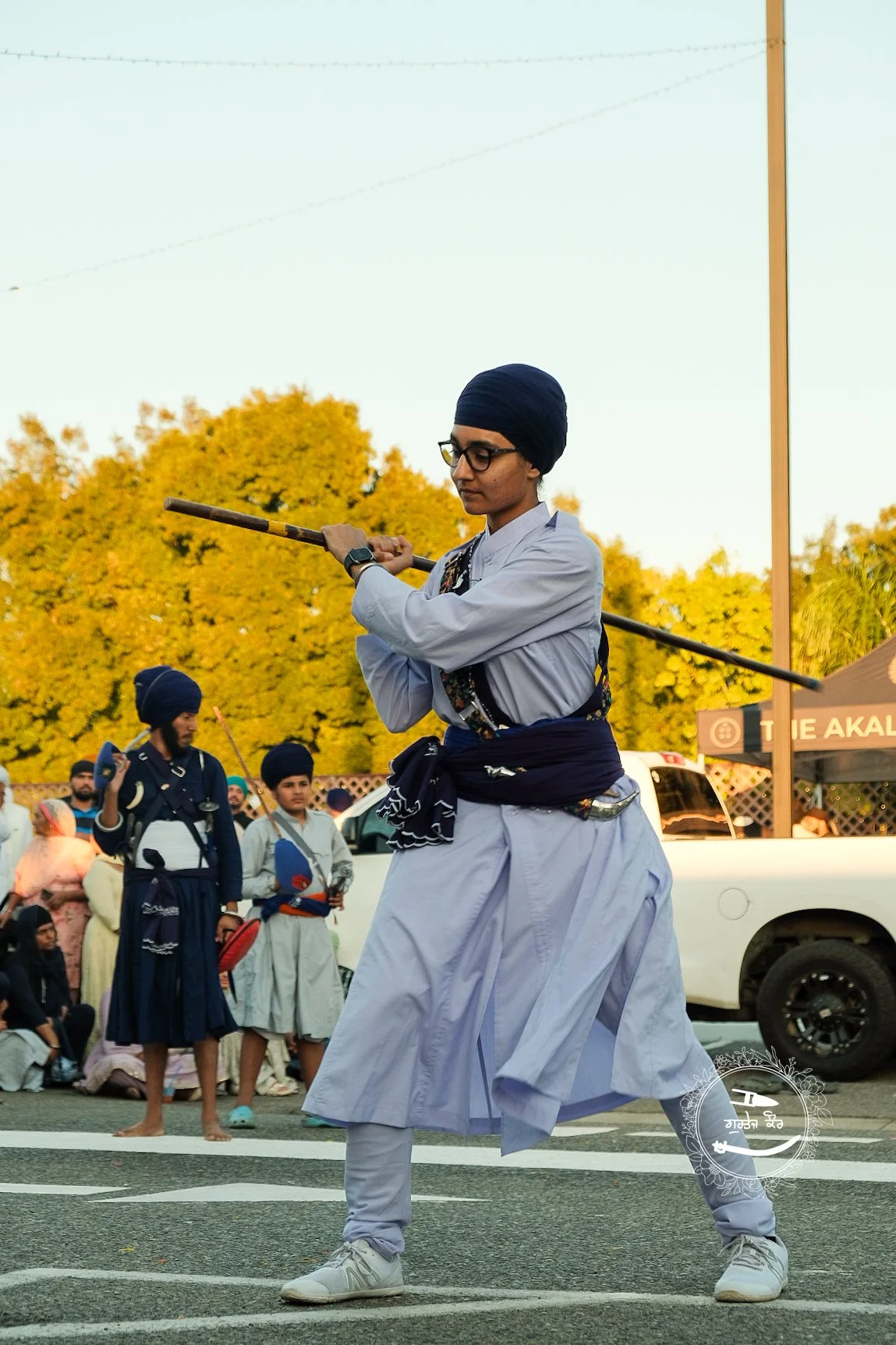 A person wearing a blue turban and glasses performs a martial arts move with a staff during a cultural event, with other participants in similar attire and a crowd in the background.
