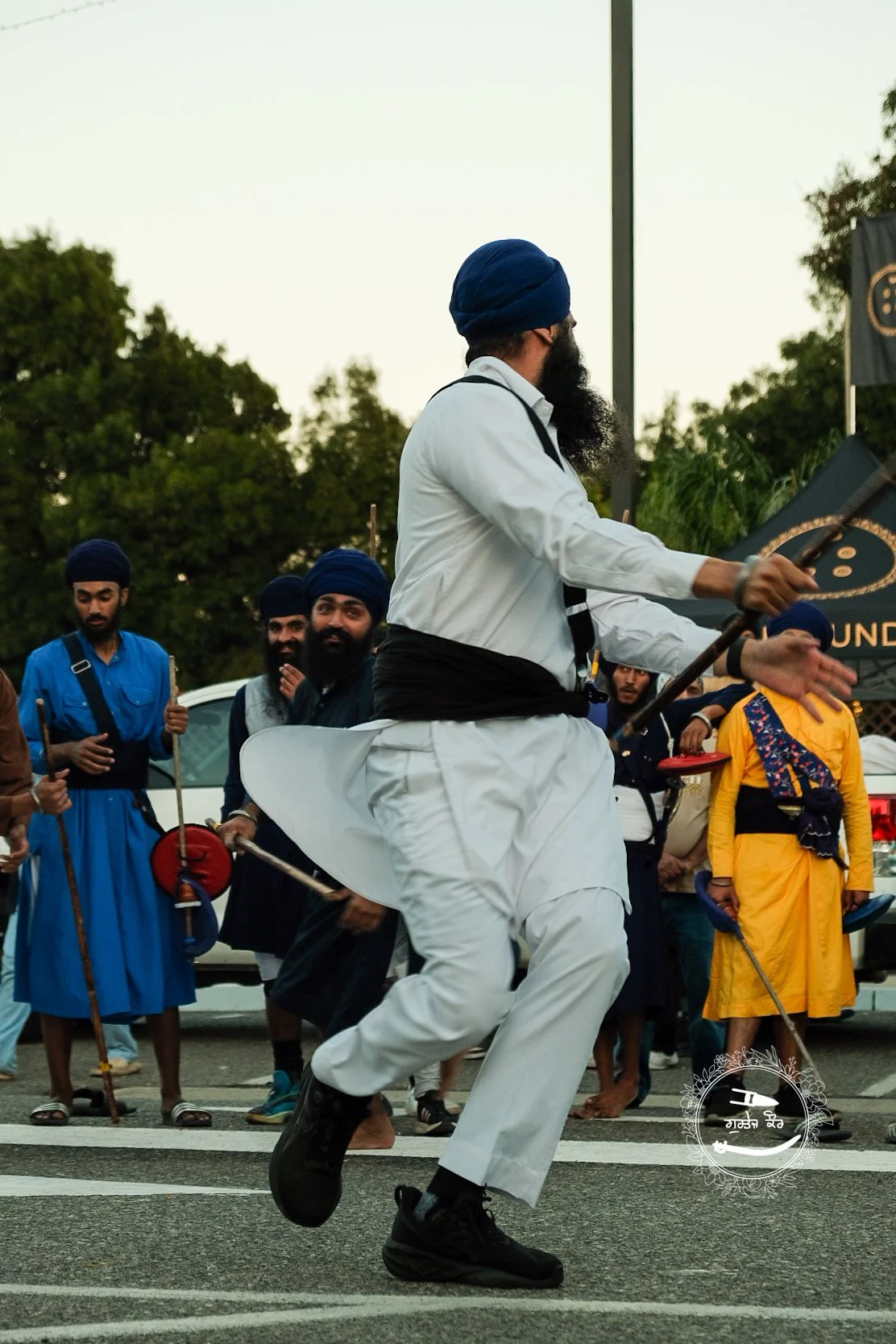 A group of people, many wearing traditional Sikh attire and turbans, participating in a cultural dance or procession outdoors. The central figure is dressed in white and appears to be leading the dance.