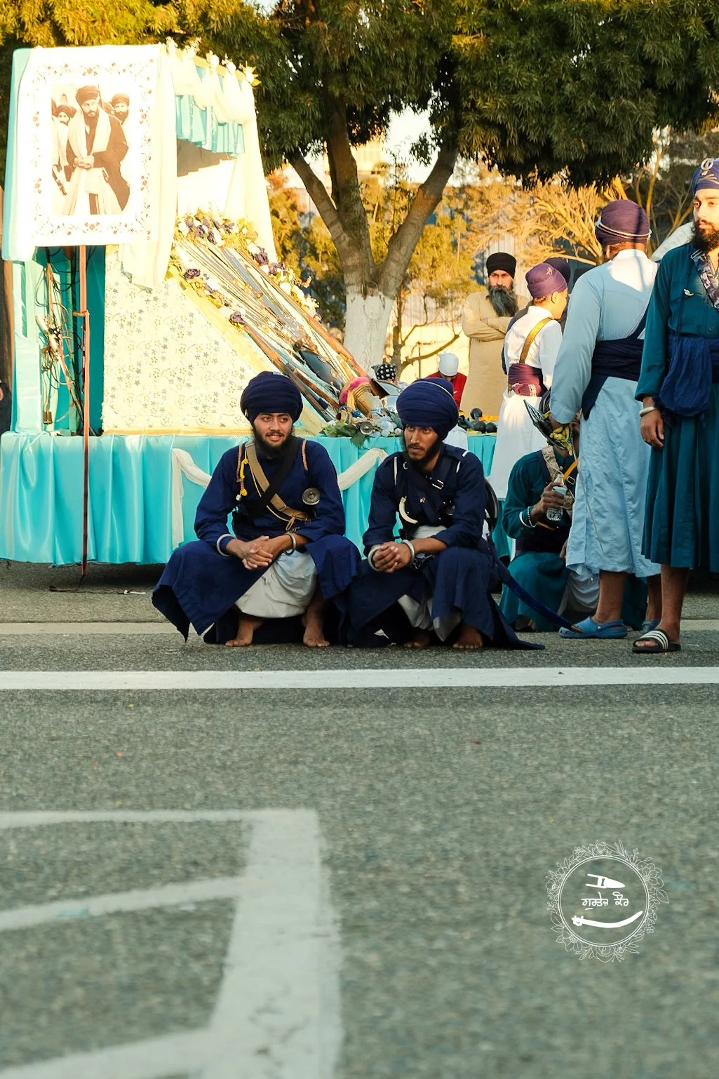 Two men sitting on the ground in traditional Sikh attire, including blue turbans and kurtas, in front of a decorated float with a religious picture and flowers, during a parade or celebration. Trees and other people in traditional clothing are visibl