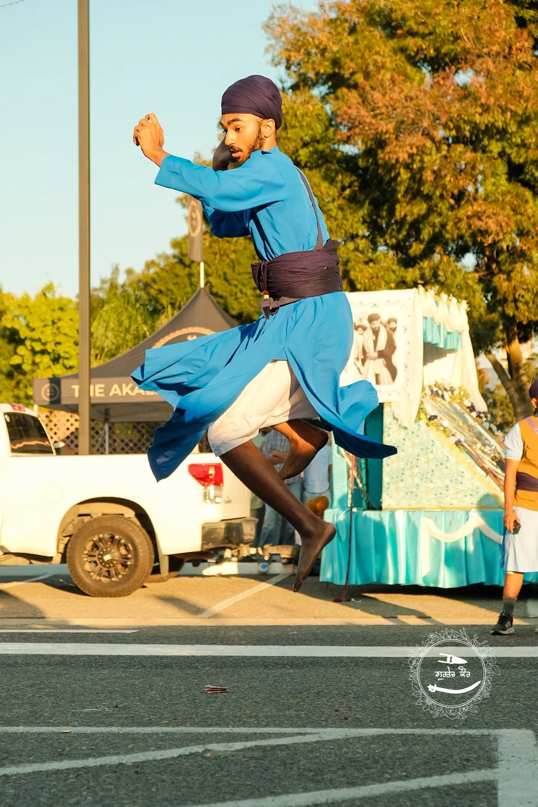 A man in traditional blue and white Indian attire, including a turban, is mid-air performing a jump during a cultural festival on a street with trees and booths in the background.