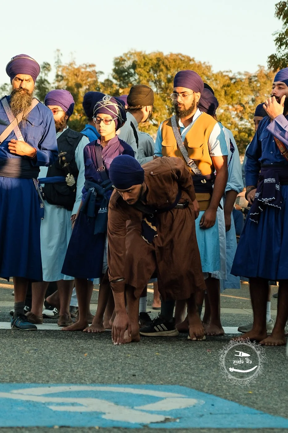 A group of Sikh men and women, some wearing turbans, gathered outdoors during daylight. One man in the foreground bends over touching the ground with his hand and foot, indicating movement or a cultural gesture. The background features trees with aut