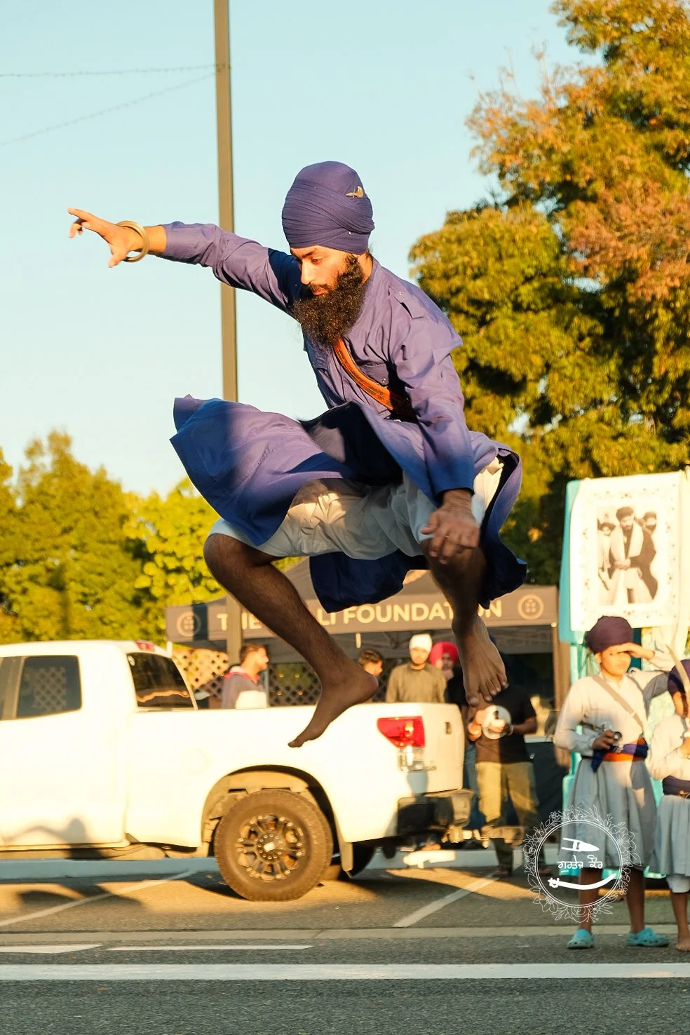 A man wearing traditional Indian attire, including a turban, is mid-air performing a jump or dance move on a street. There are onlookers and trees in the background.