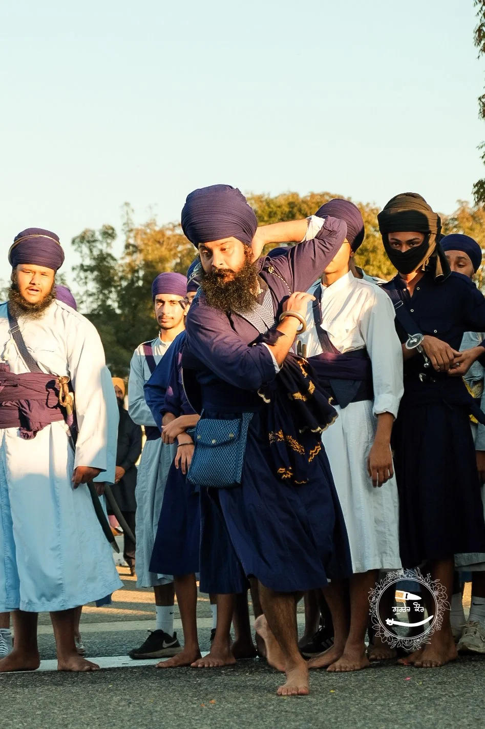 Group of Sikh men dressed in traditional attire, gathering outdoors during daytime, with some wearing turbans and others with head coverings, and one man carrying a bag.