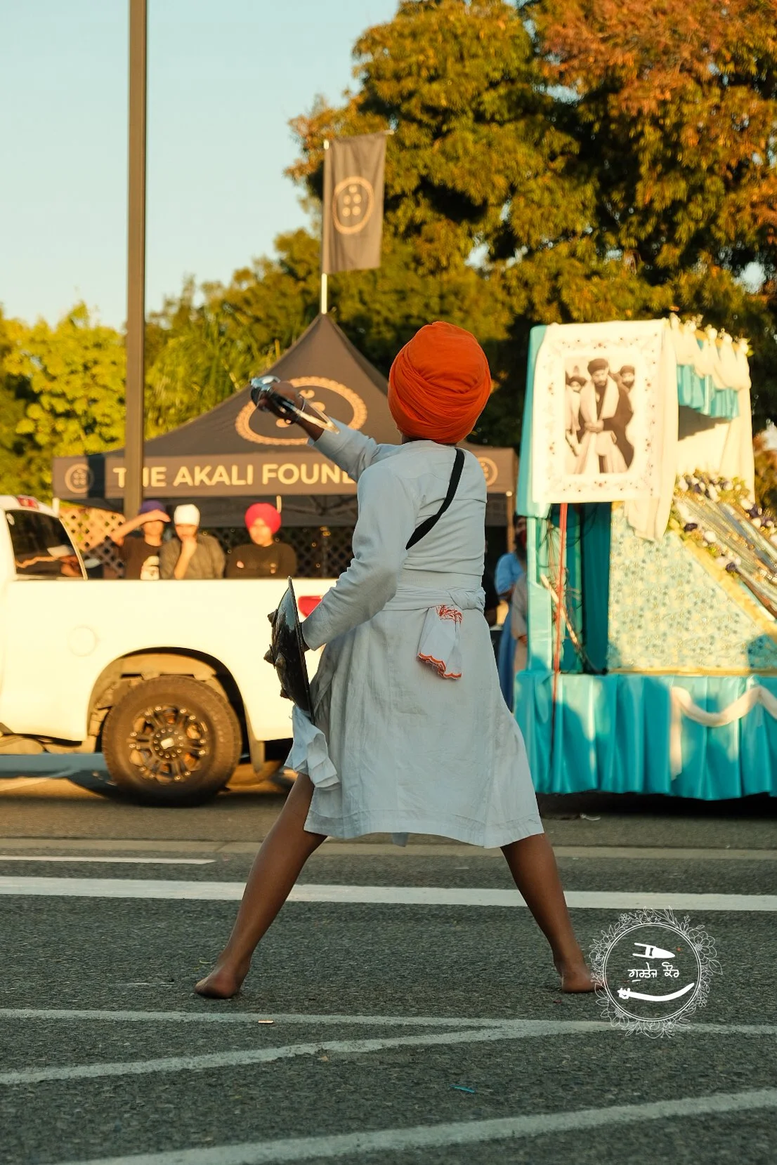 A person wearing a white dress and an orange turban appears to be dancing or performing in the middle of a street, with a cap in hand, in front of a decorated float and a tent with people and a sign that reads "The Akali Foundation."