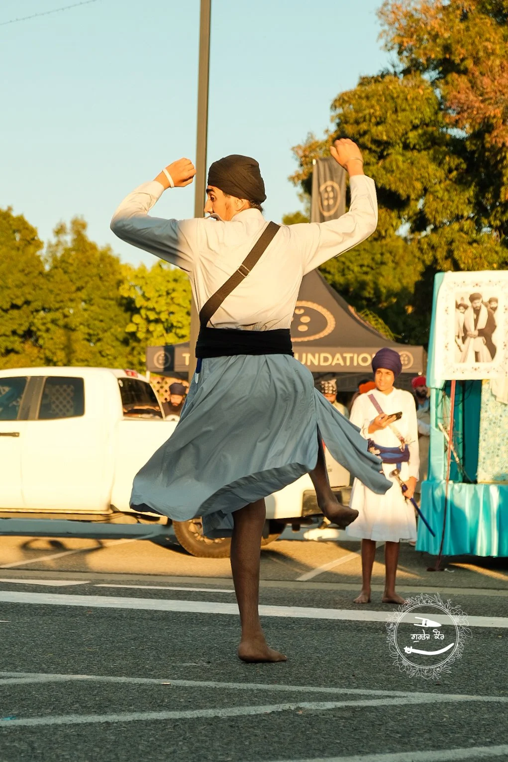 A man dressed in traditional Indian attire, including a turban and long skirt, is dancing barefoot on a street during a cultural event, with others watching in the background.