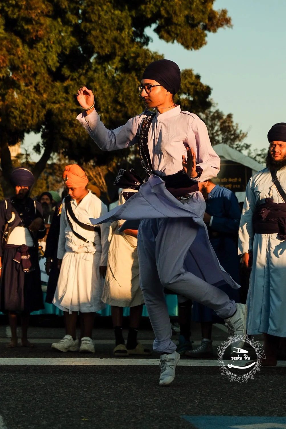 A person in traditional Punjabi attire, including a turban and glasses, is dancing in a crowd during a cultural event. Others in similar attire stand in the background.