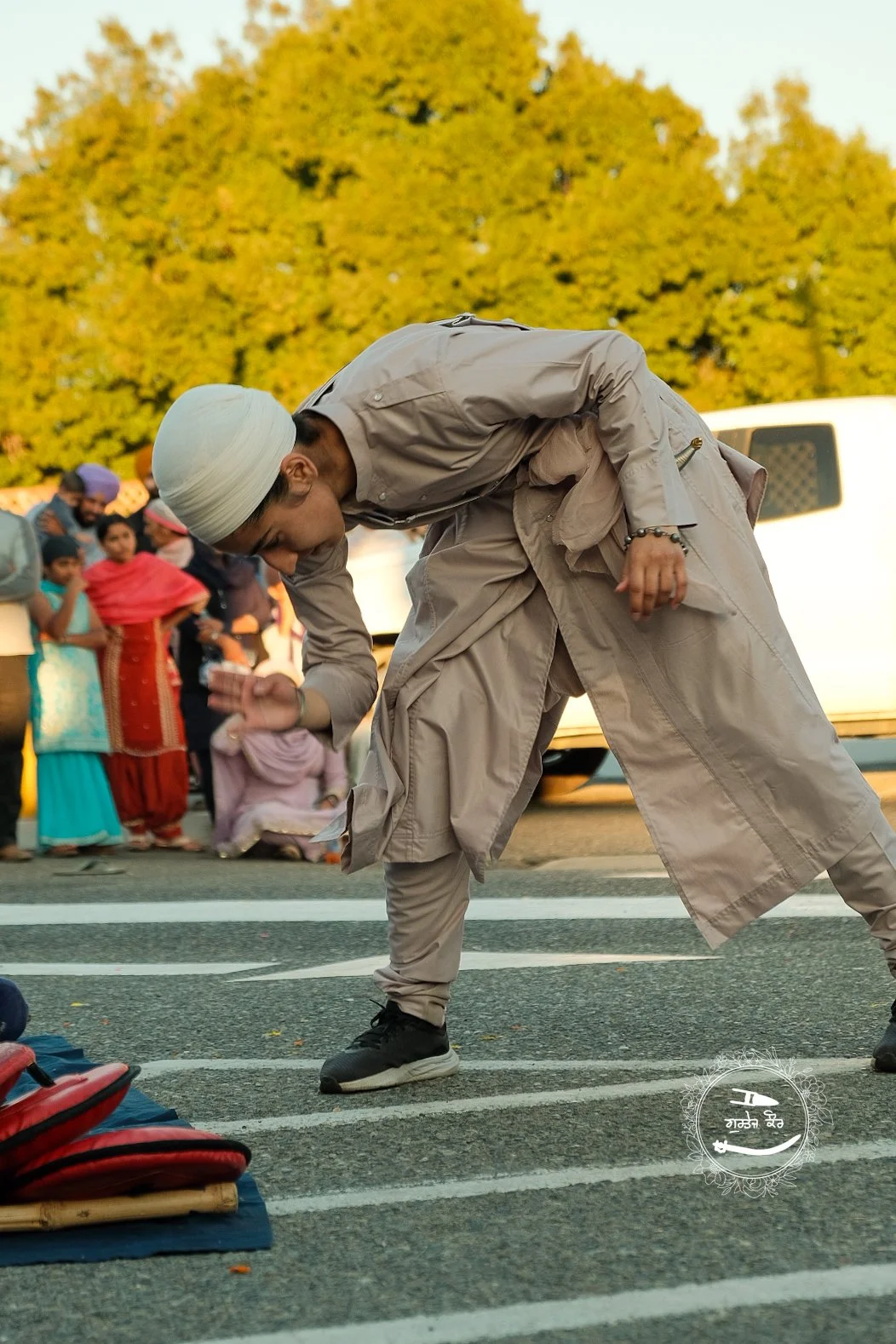 A man wearing traditional Sikh attire, including a white turban and beige clothing, bending forward on a street with a group of people, including children, in the background. The scene appears to be outdoors during the day with trees and a white vehi