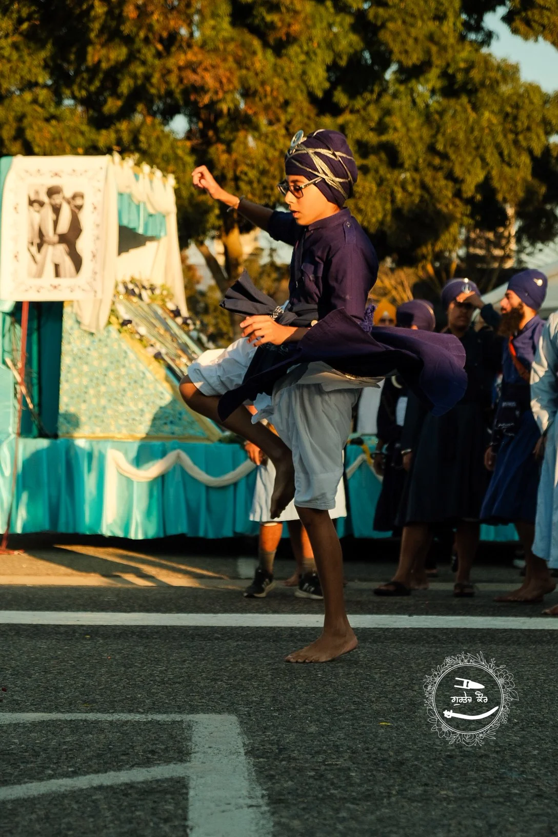 Young man dancing during a parade, dressed in traditional clothing, barefoot, with a colorful turban, holding a musical instrument, with a decorated float in the background and other participants wearing turbans.